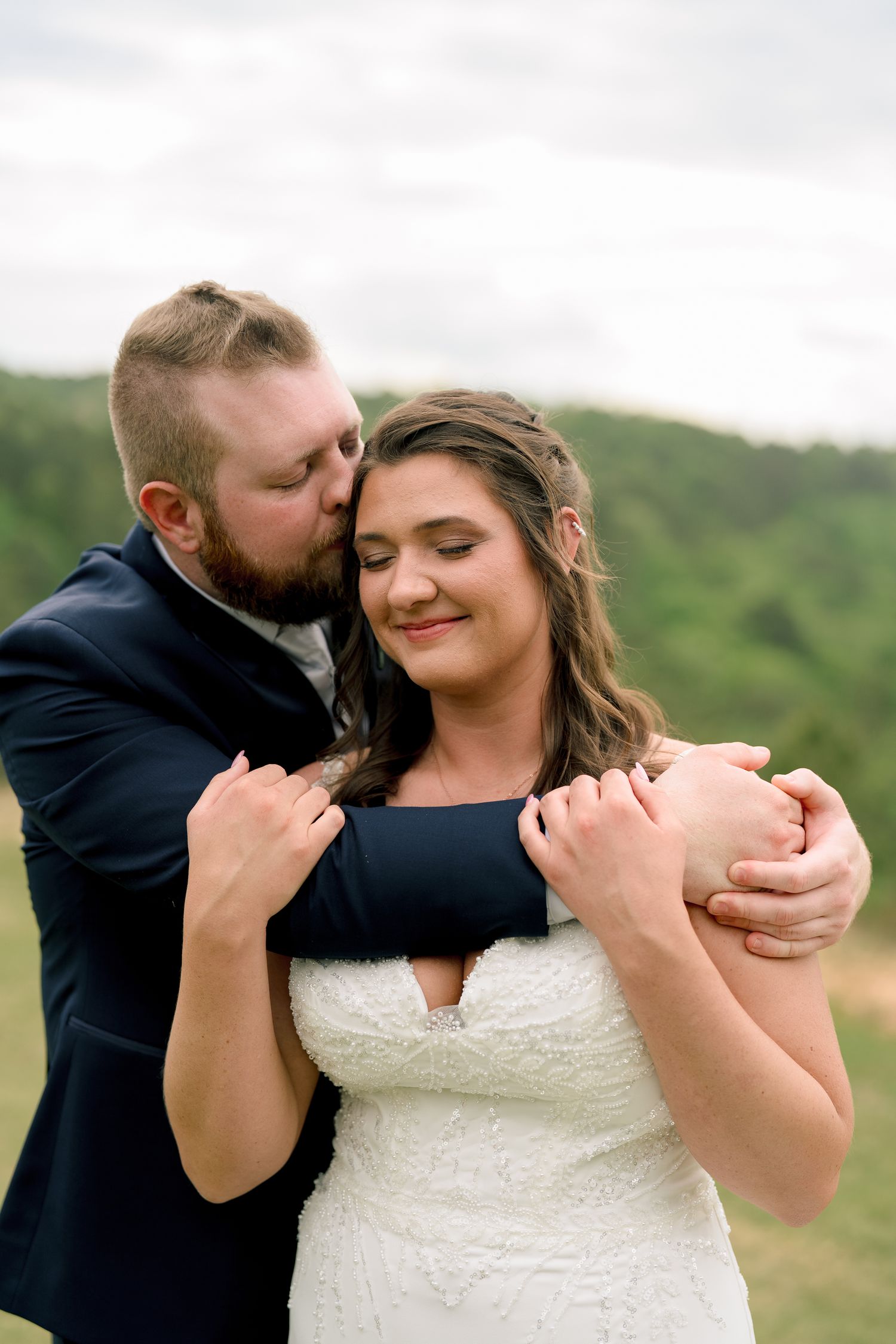 Groom in navy suit embracing bride in white lace dress, kissing her cheek outdoors with green hills behind.