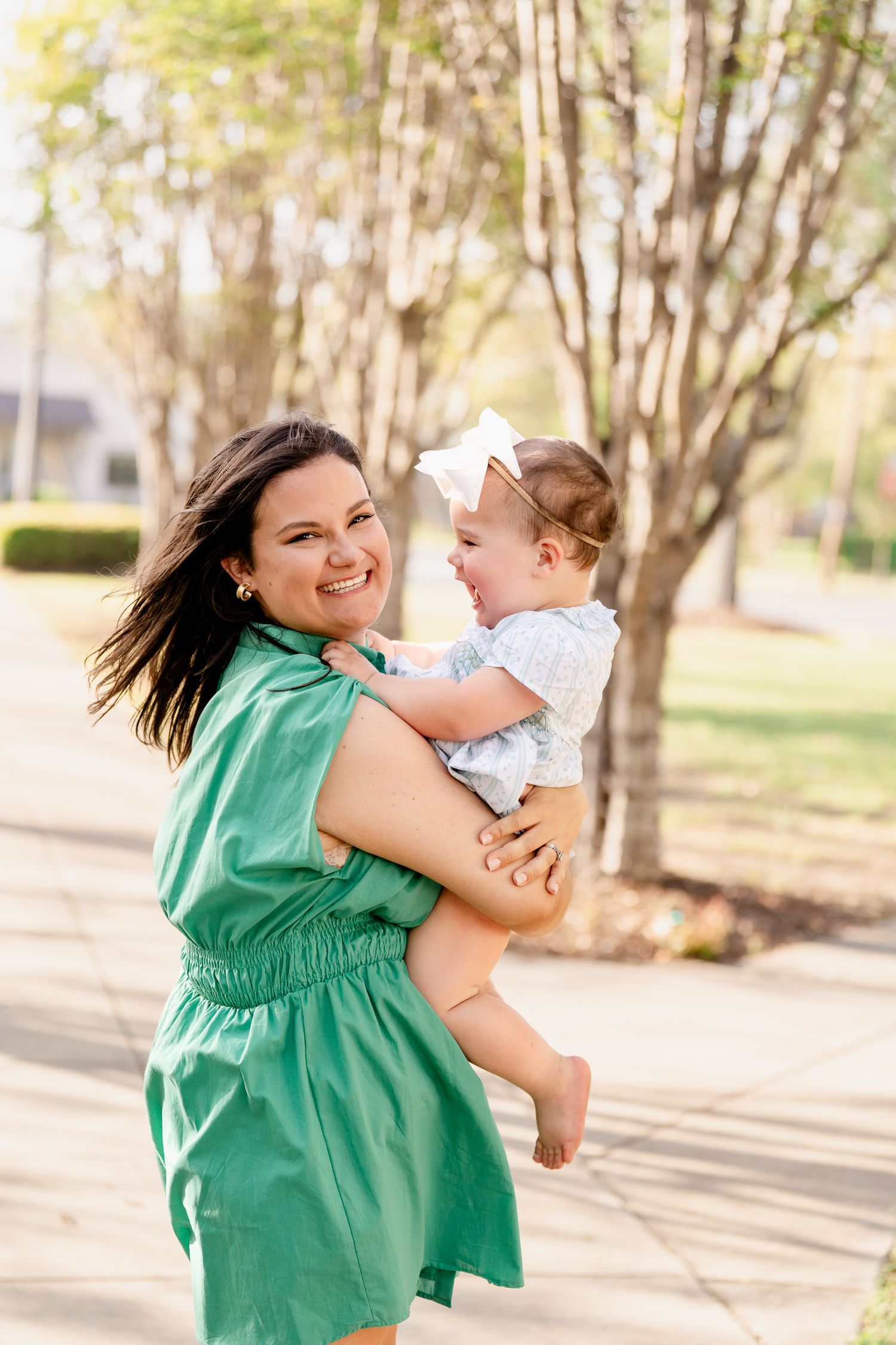 A woman in a green dress holds and laughs with a baby outdoors among trees.