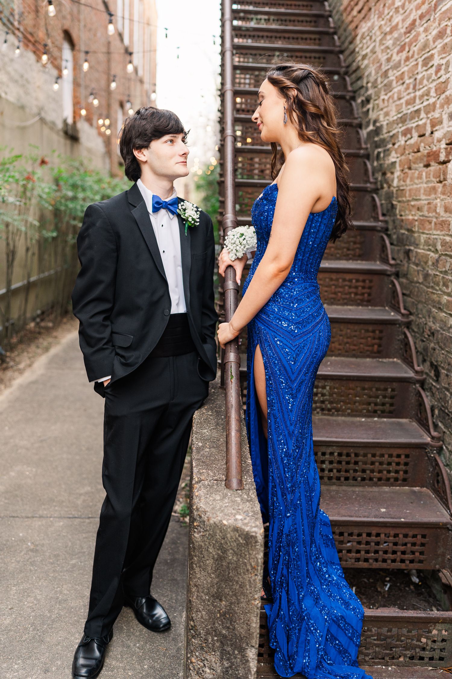 Couple in formal attire poses by rustic brick wall and metal staircase.