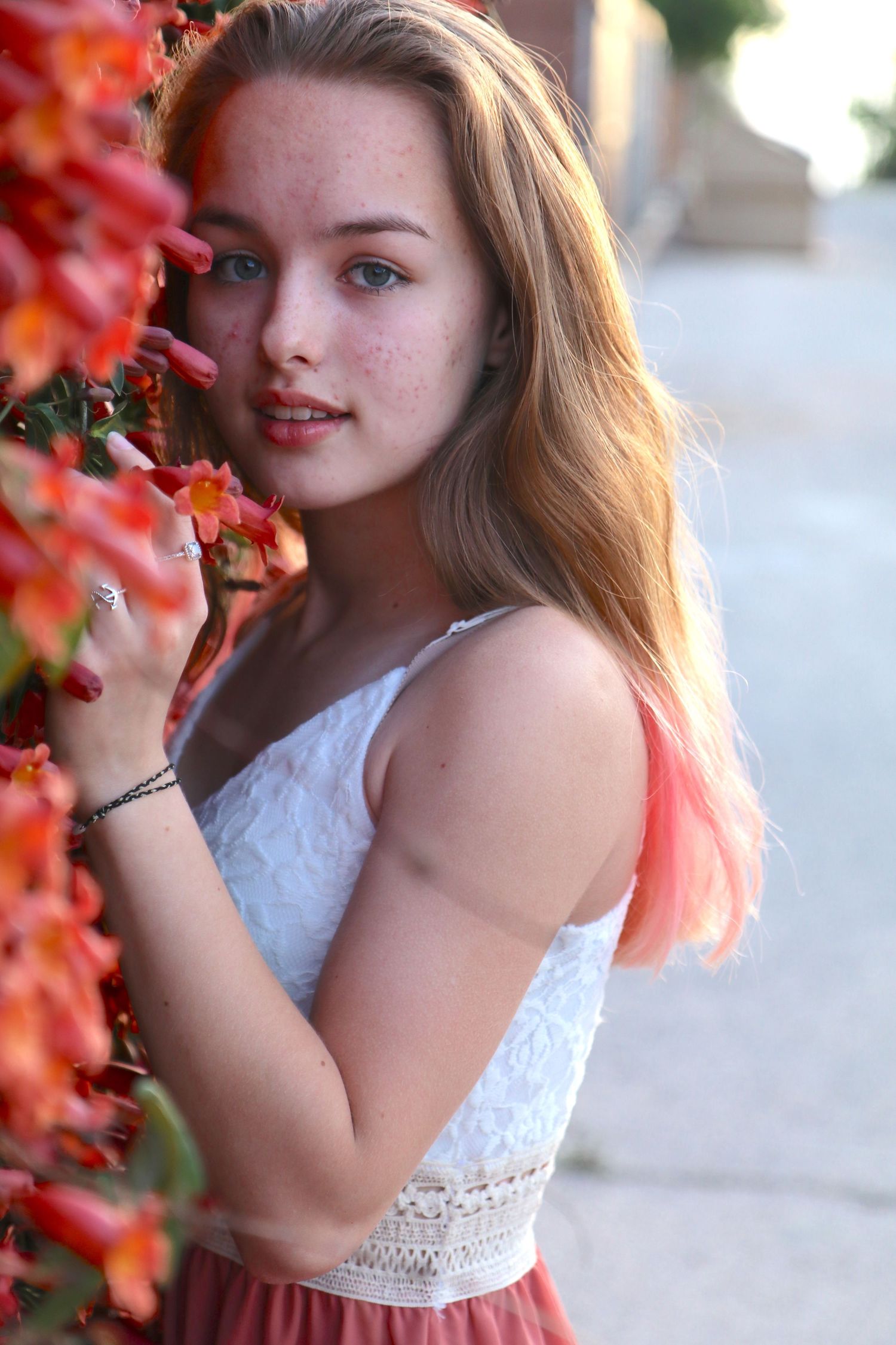 Young child in a white lace dress poses near vibrant red flowers in a garden setting.