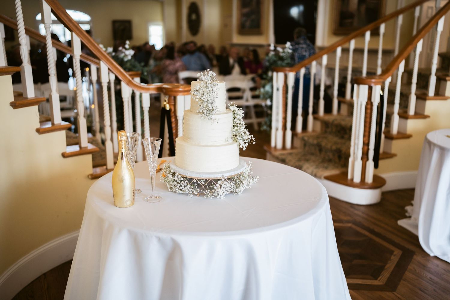 Elegant white wedding cake displayed on round table with staircase in background.