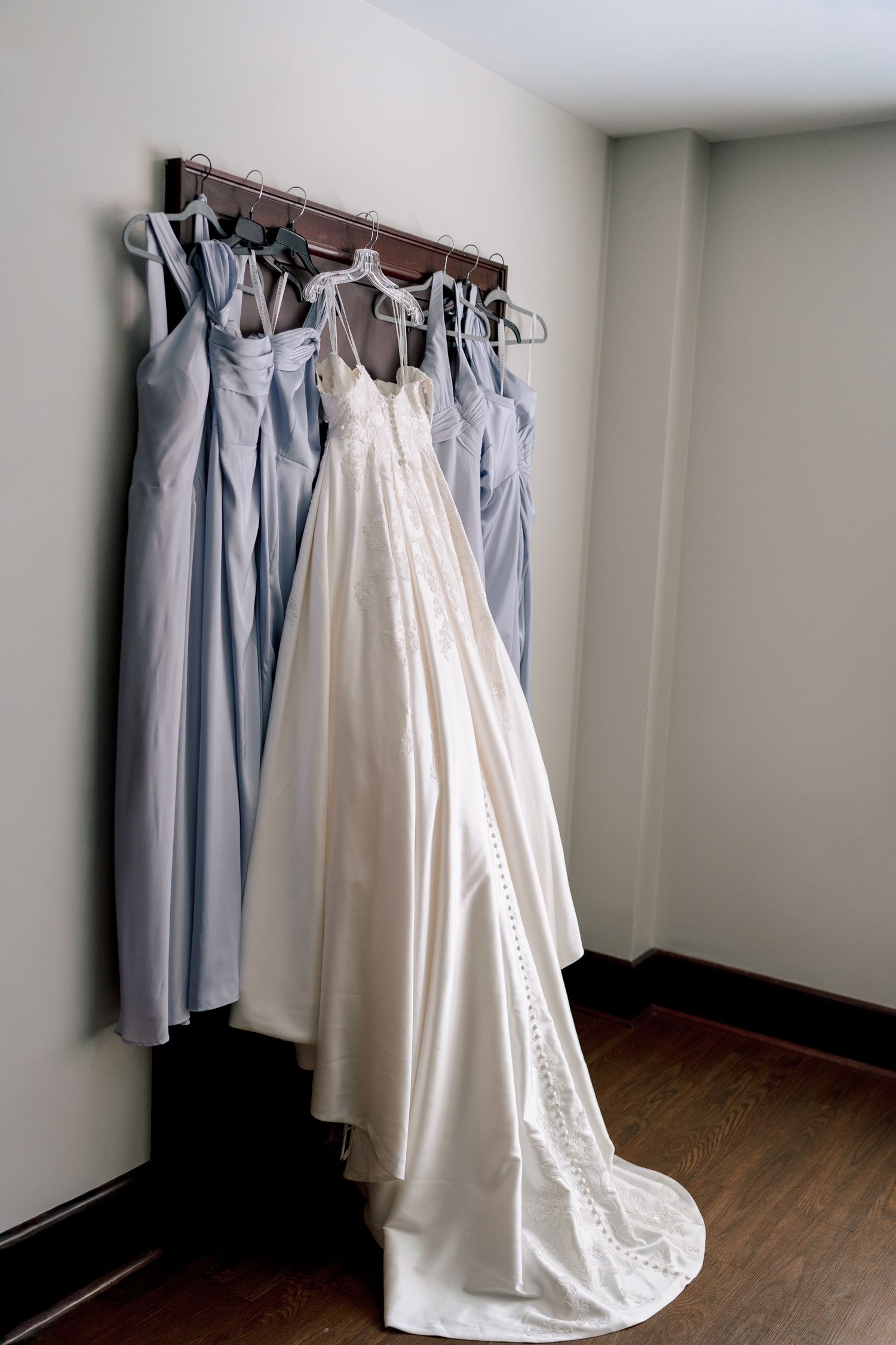 A series of light blue bridesmaid dresses and one white wedding gown hanging on a clothing rack against a white wall.