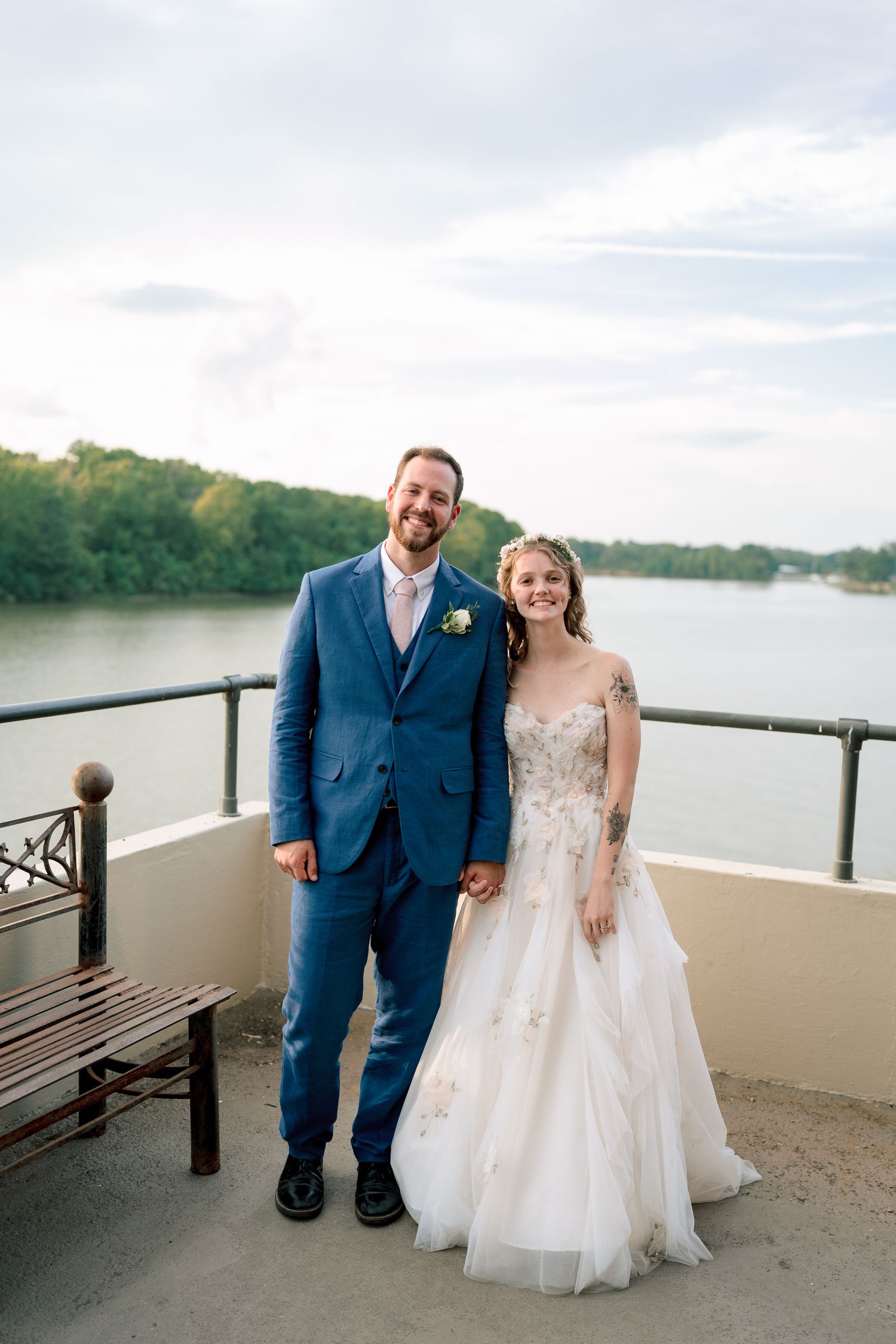 Smiling bride and groom pose on waterfront terrace, she in floral gown, he in blue suit with boutonniere.
