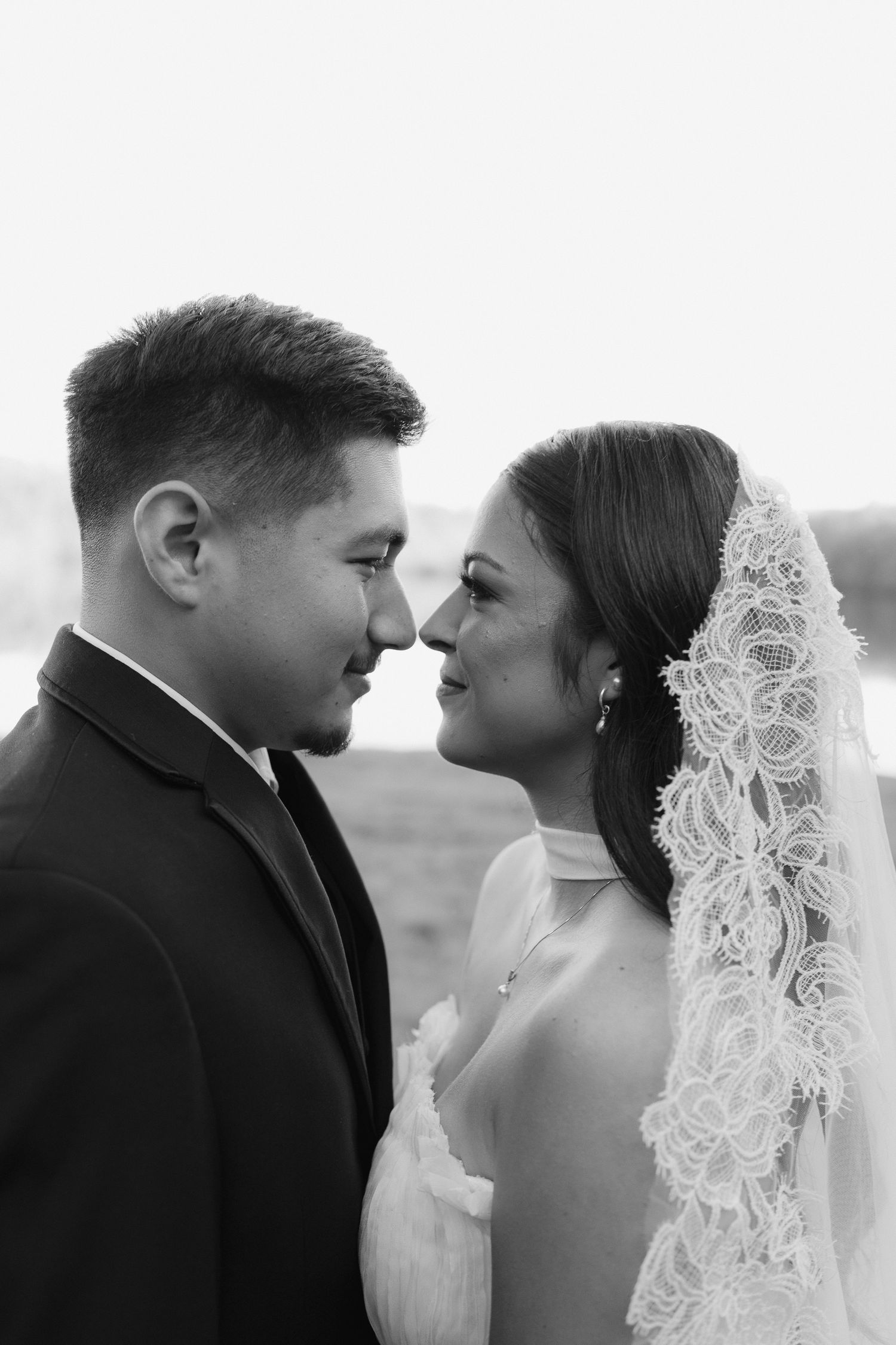 Black and white portrait of newlywed couple in formal attire, bride wearing lace veil, sharing an intimate moment.