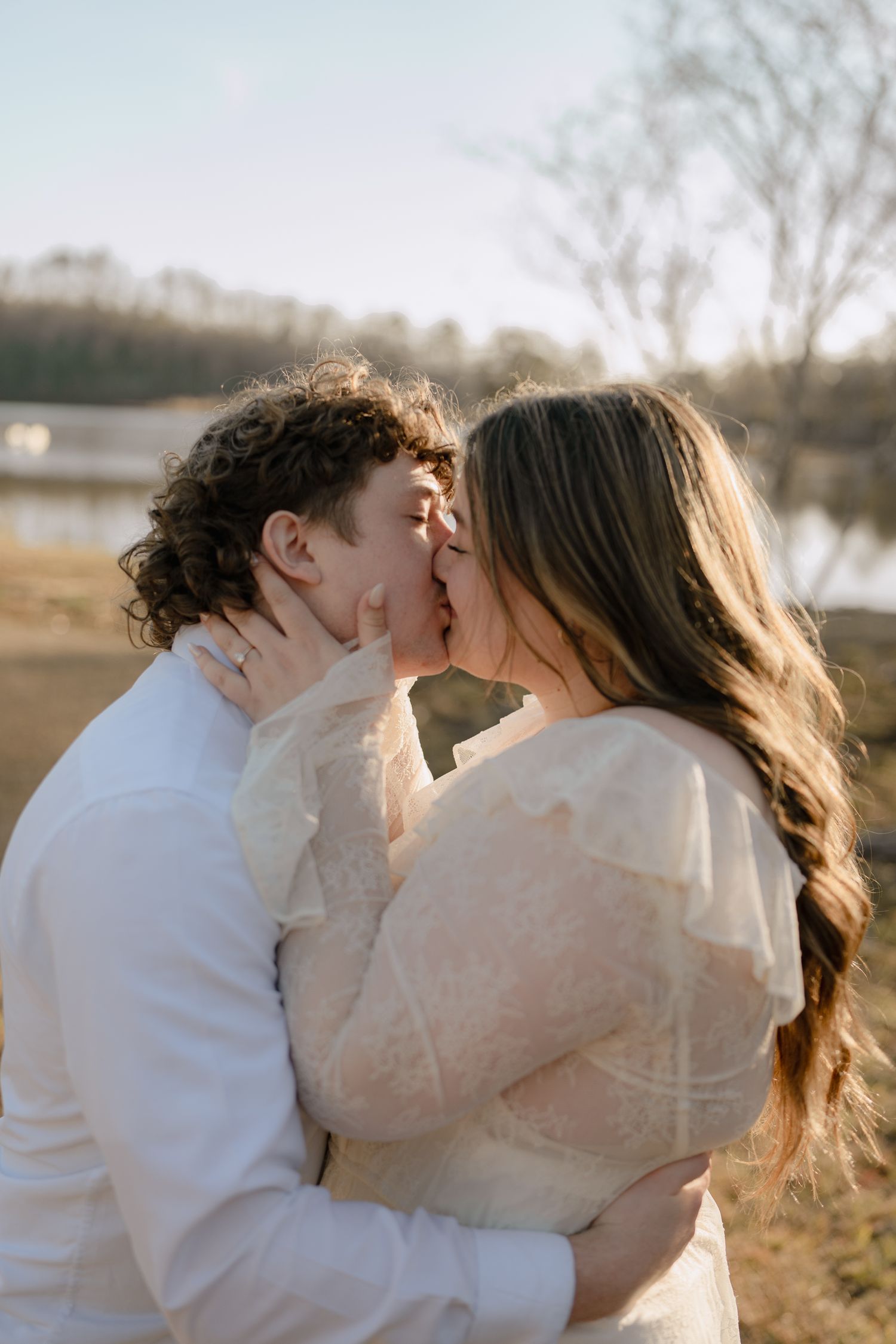 Couple sharing a romantic kiss outdoors near a lake in soft golden light.