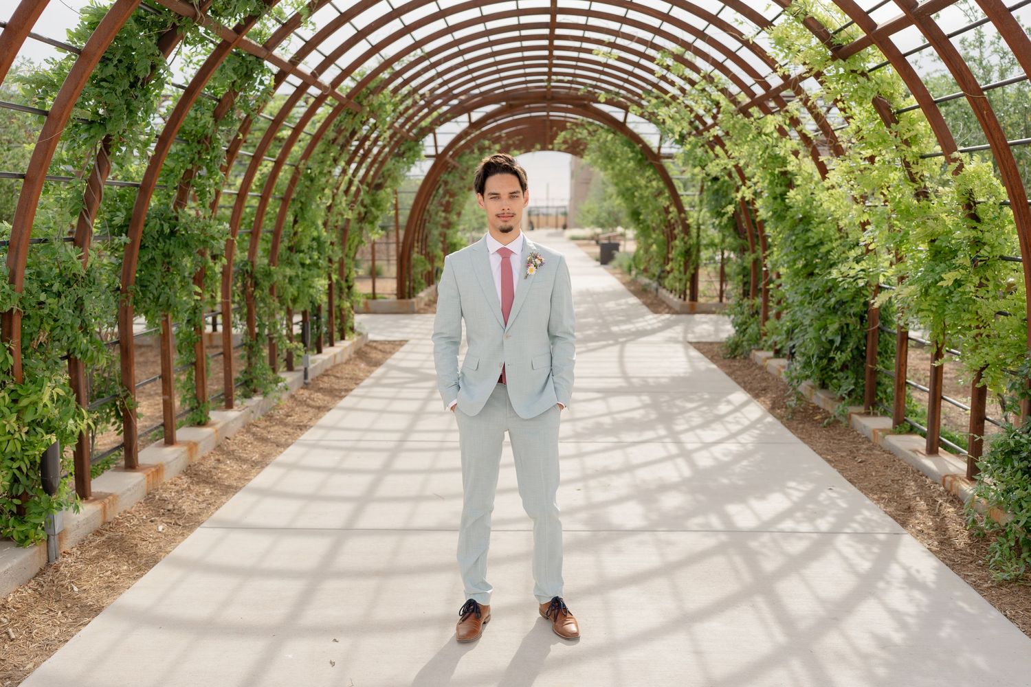 Wedding party poses in a curved garden archway with greenery and rustic wooden beams overhead.