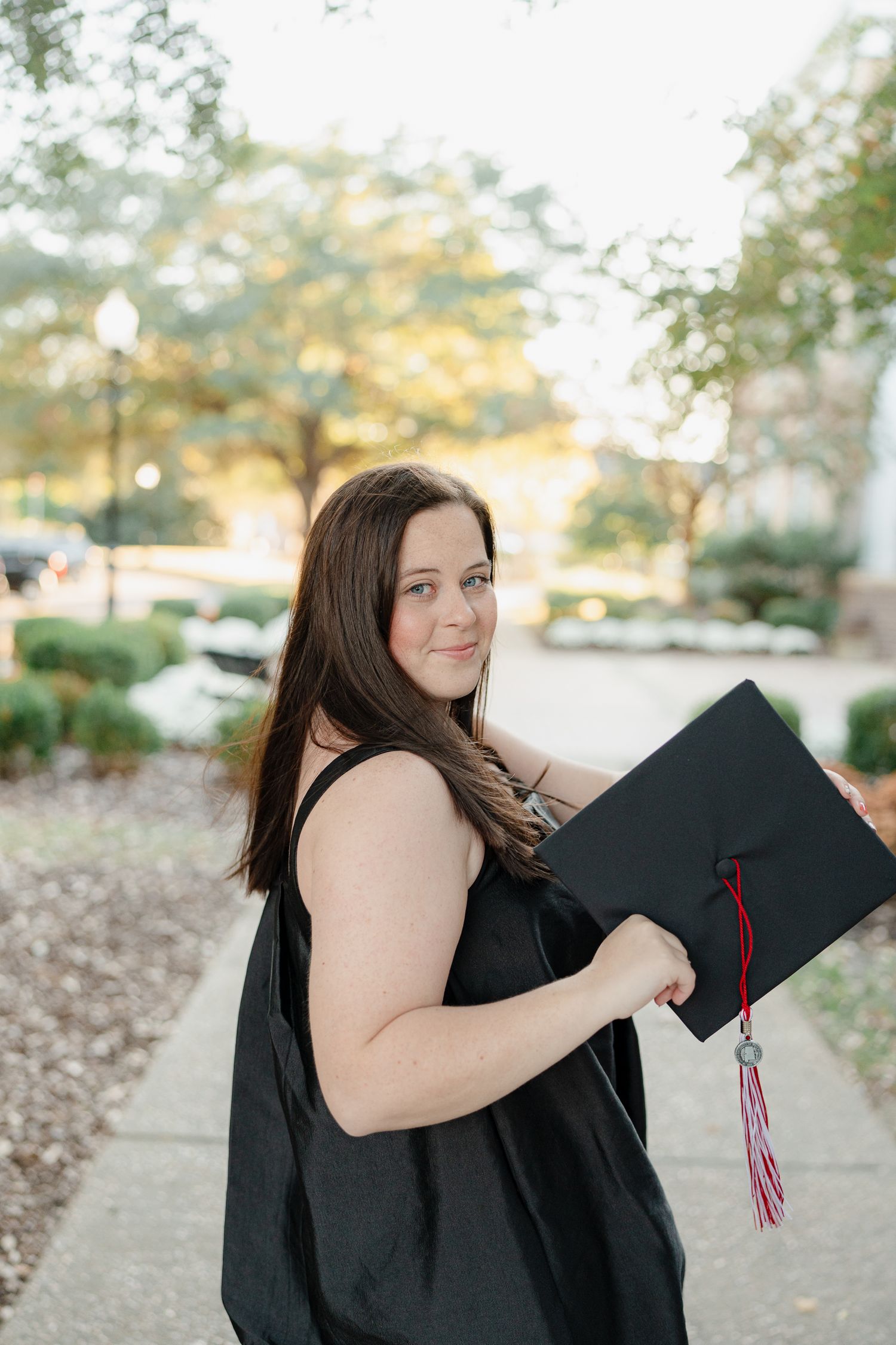Graduate in black cap and gown poses with diploma during golden hour outdoor photo session.