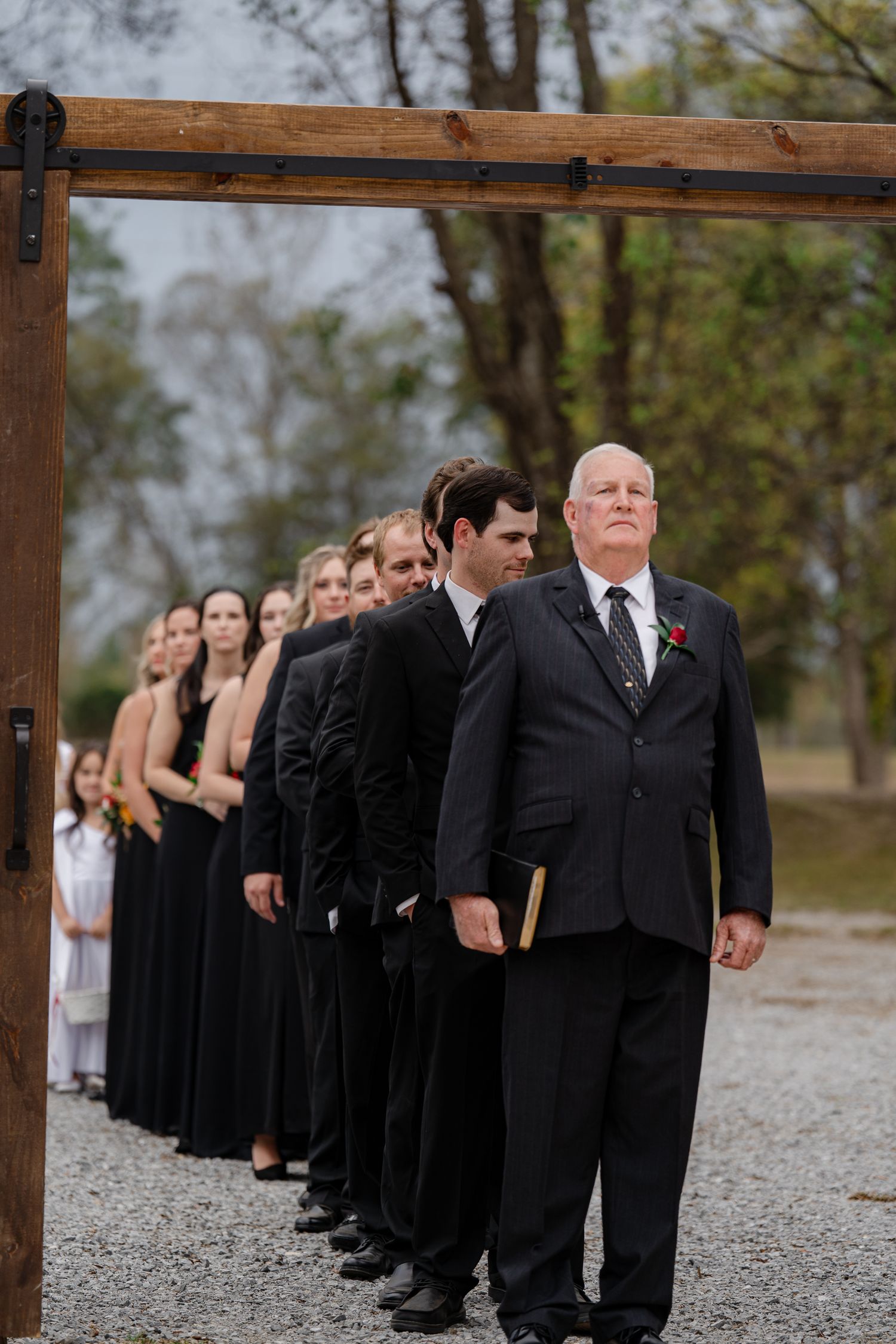 Wedding party members lined up under a wooden arch during an outdoor ceremony.