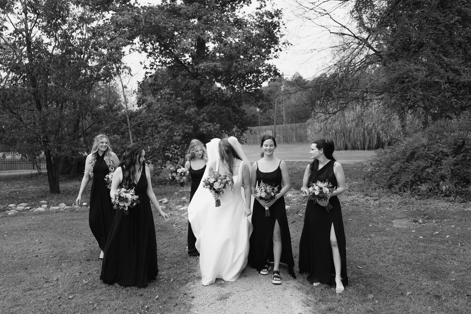 Black and white photo of bridesmaids in long black dresses walking with bride through a park setting.