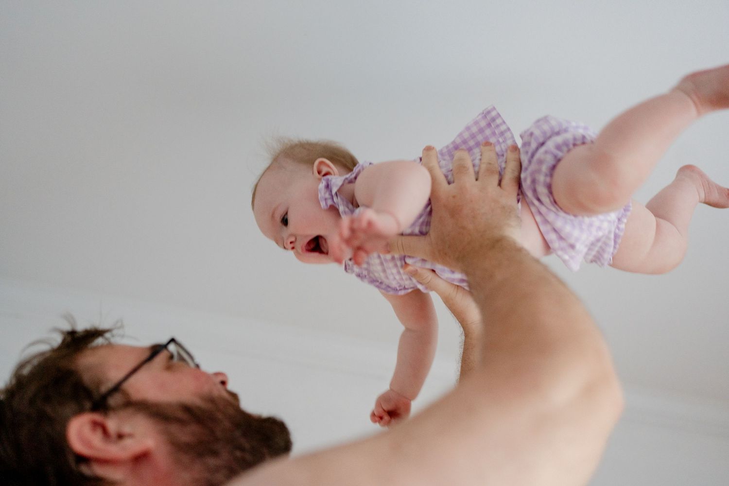 A parent lying down holds and plays with a smiling baby against a white background.