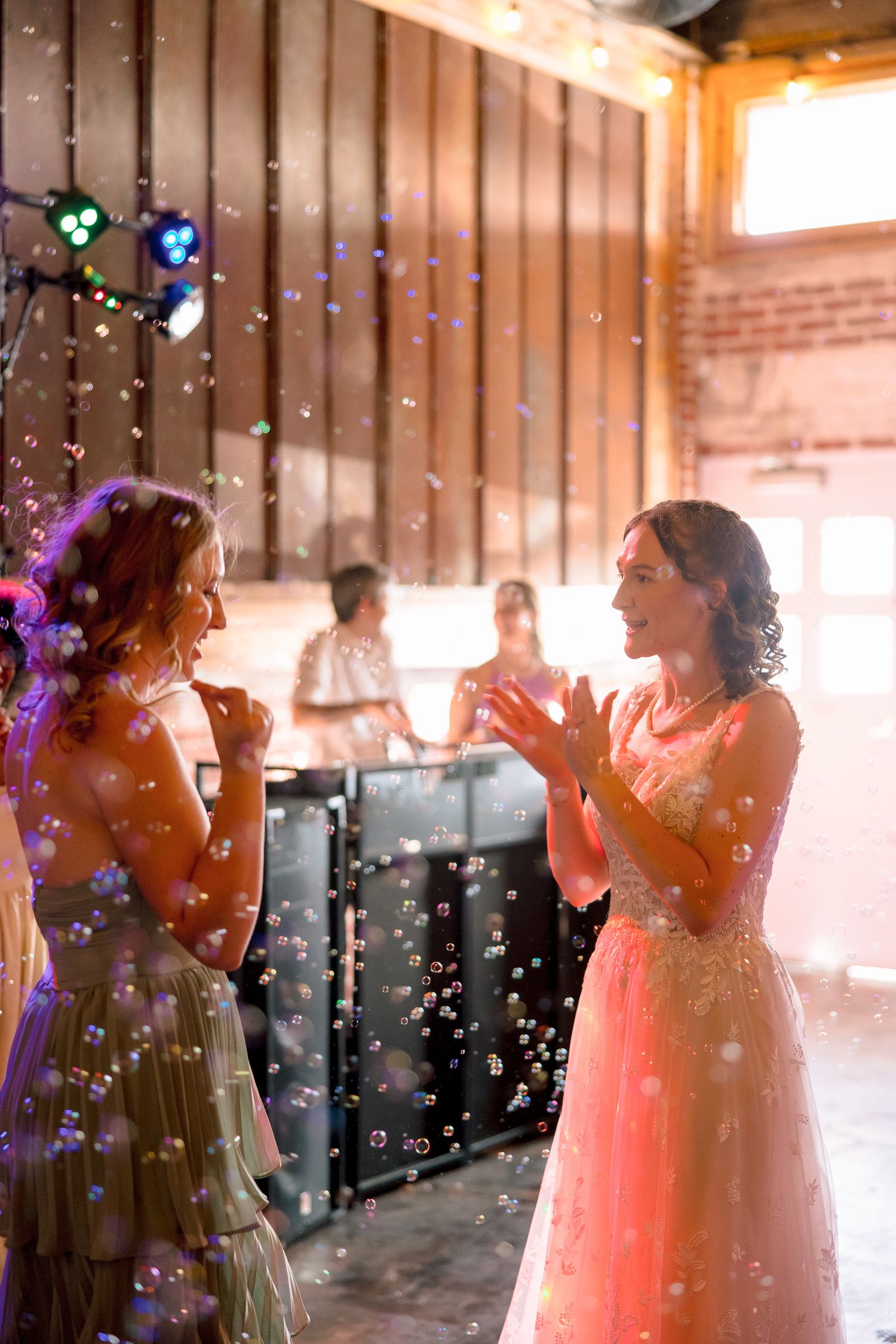 Two bridesmaids in elegant gowns laugh and play with bubbles on a warmly lit wedding reception dance floor.