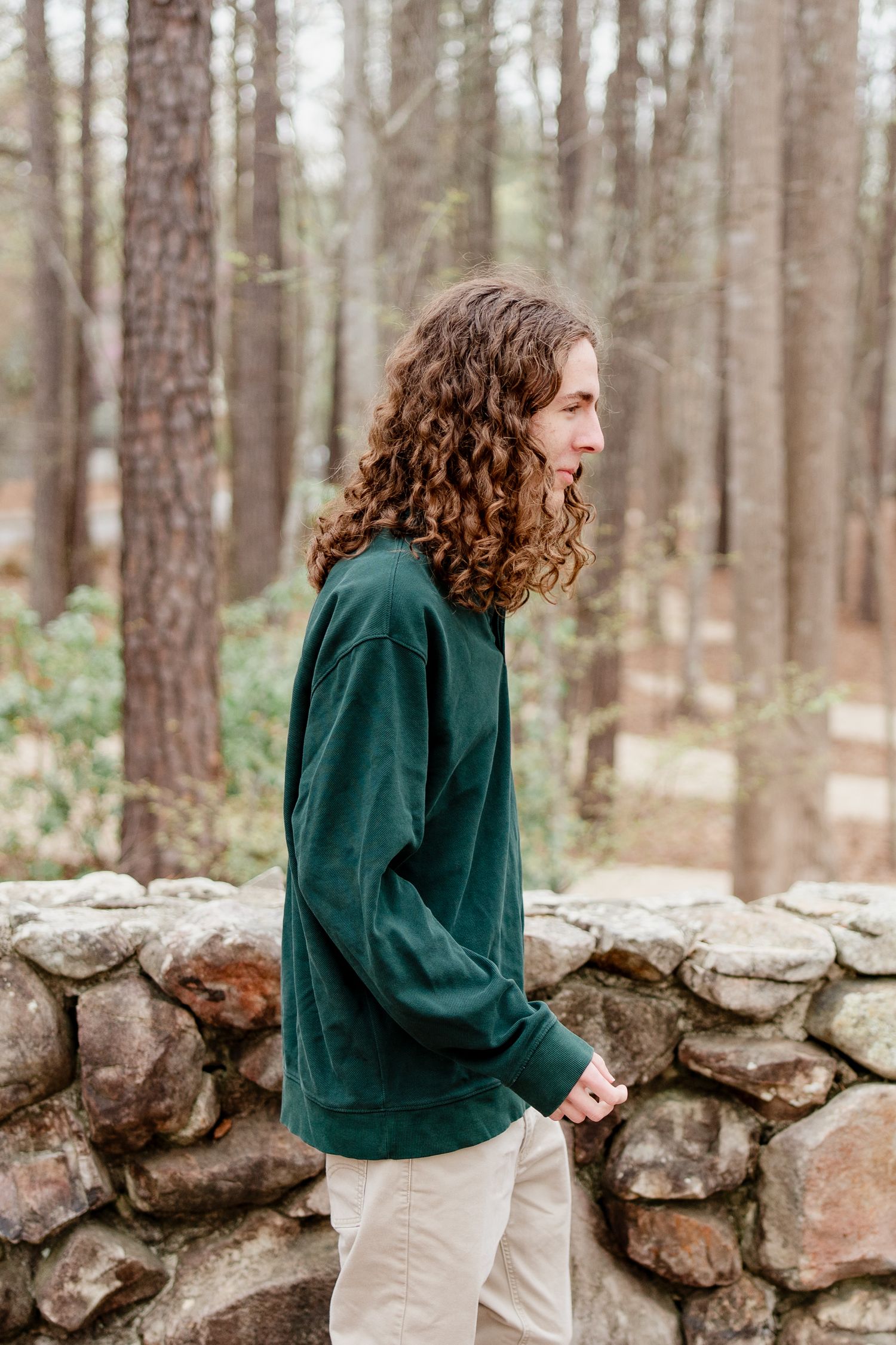 A sequence of casual poses along a stone wall in a wooded area wearing a green sweater.