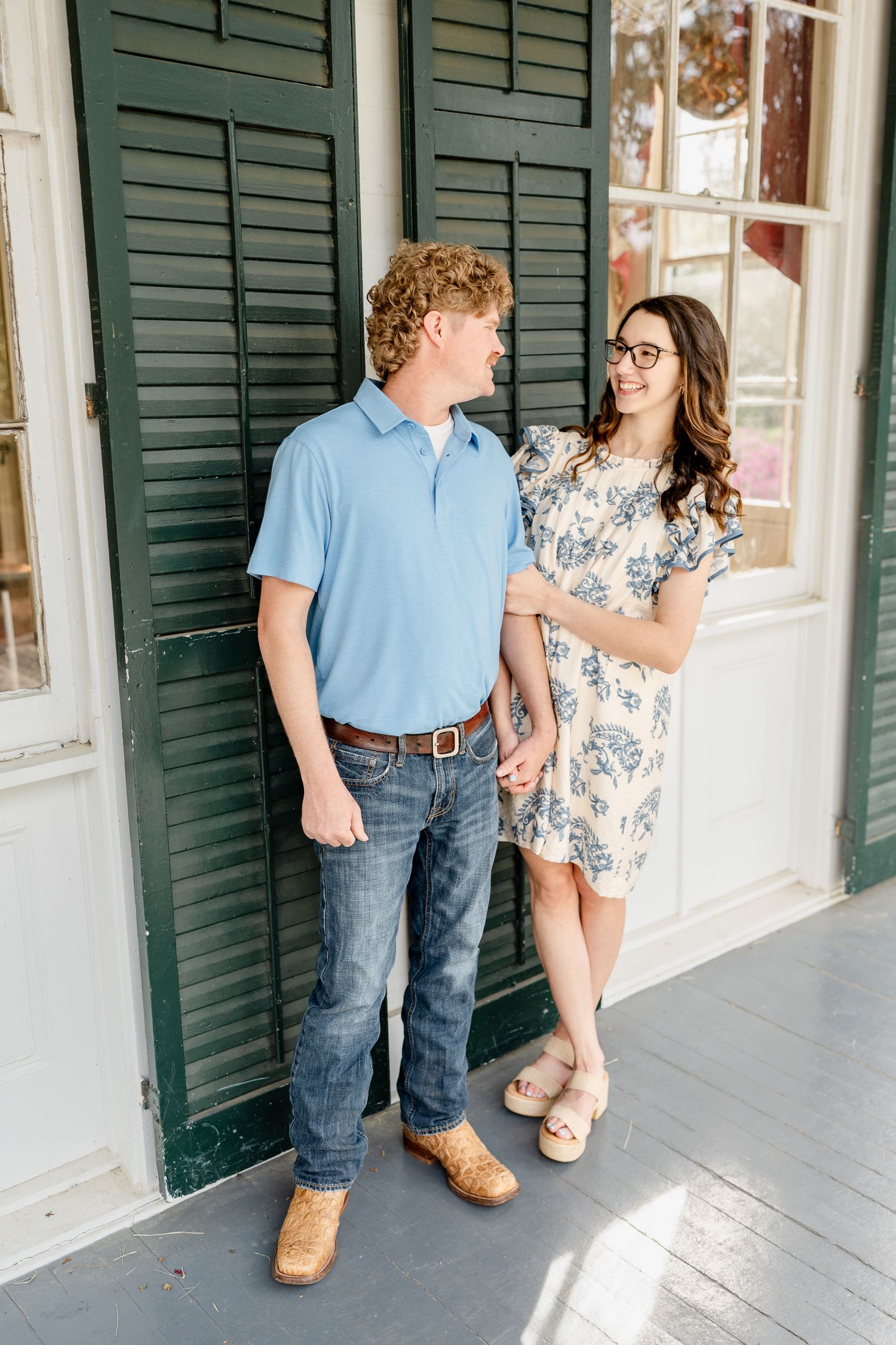 A couple stands by green shutters during a casual outdoor portrait session.