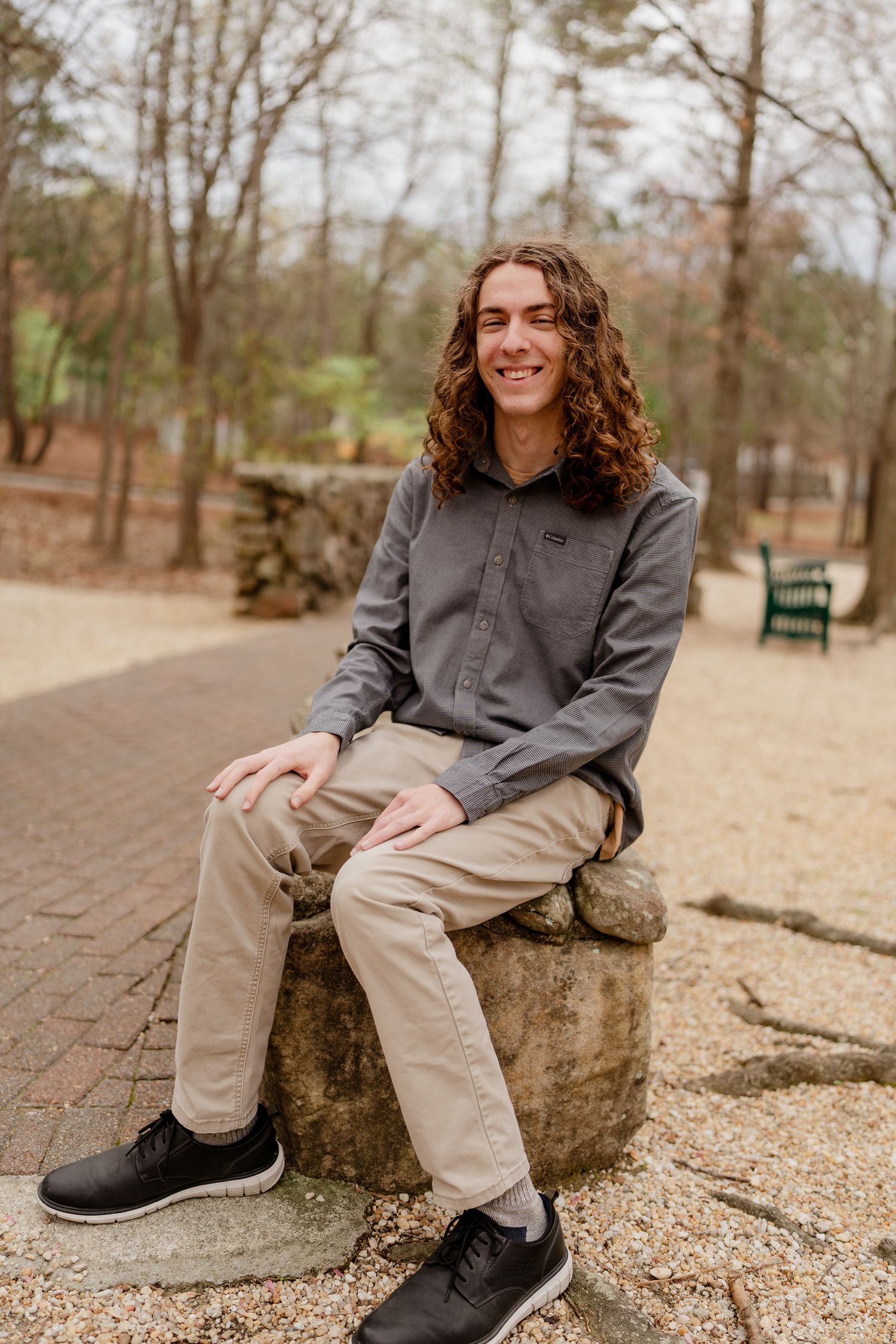 Person in grey button-down shirt sitting on large rock in park setting.