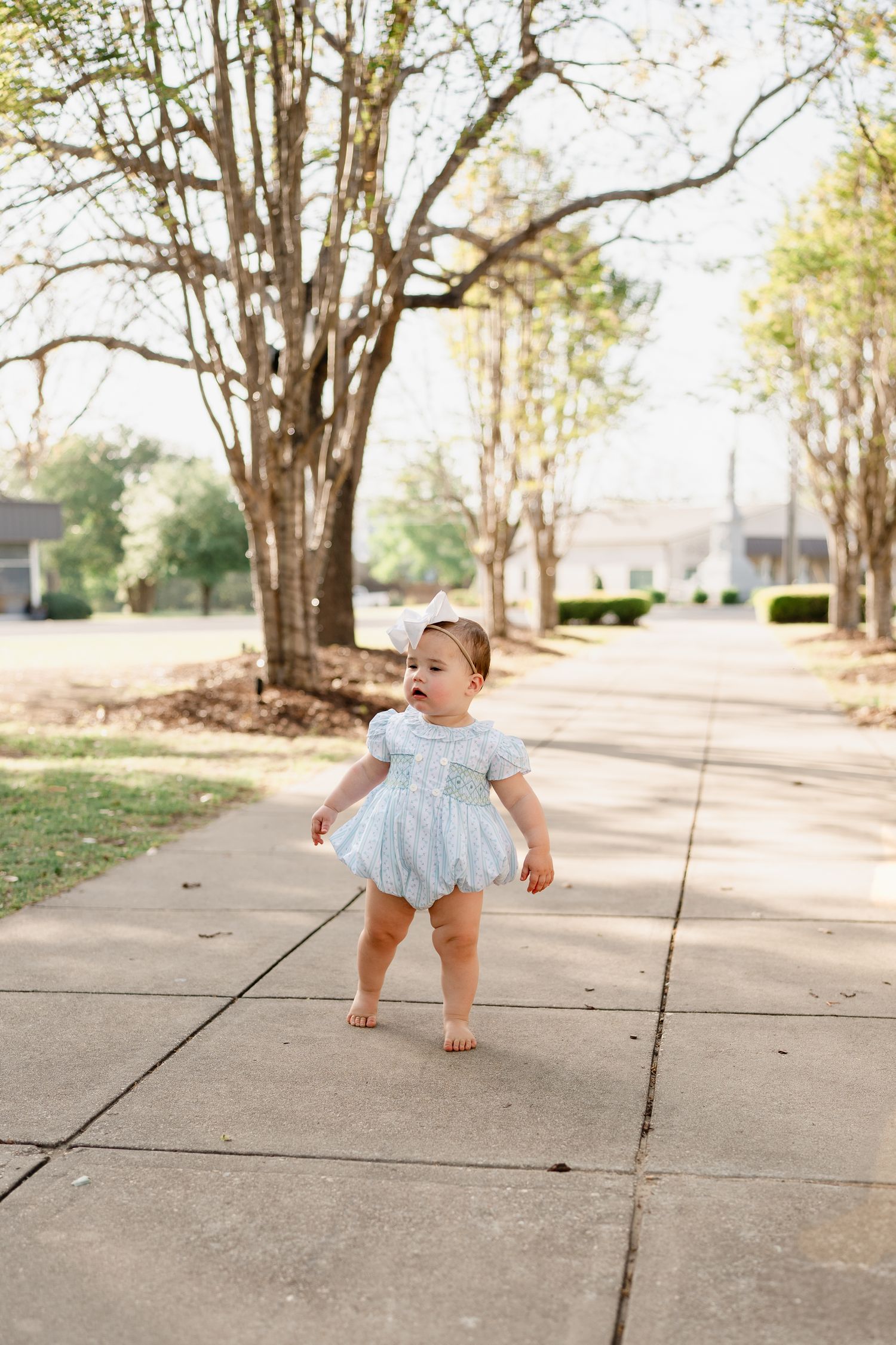 Toddler in light blue dress stands barefoot on a tree-lined sidewalk.