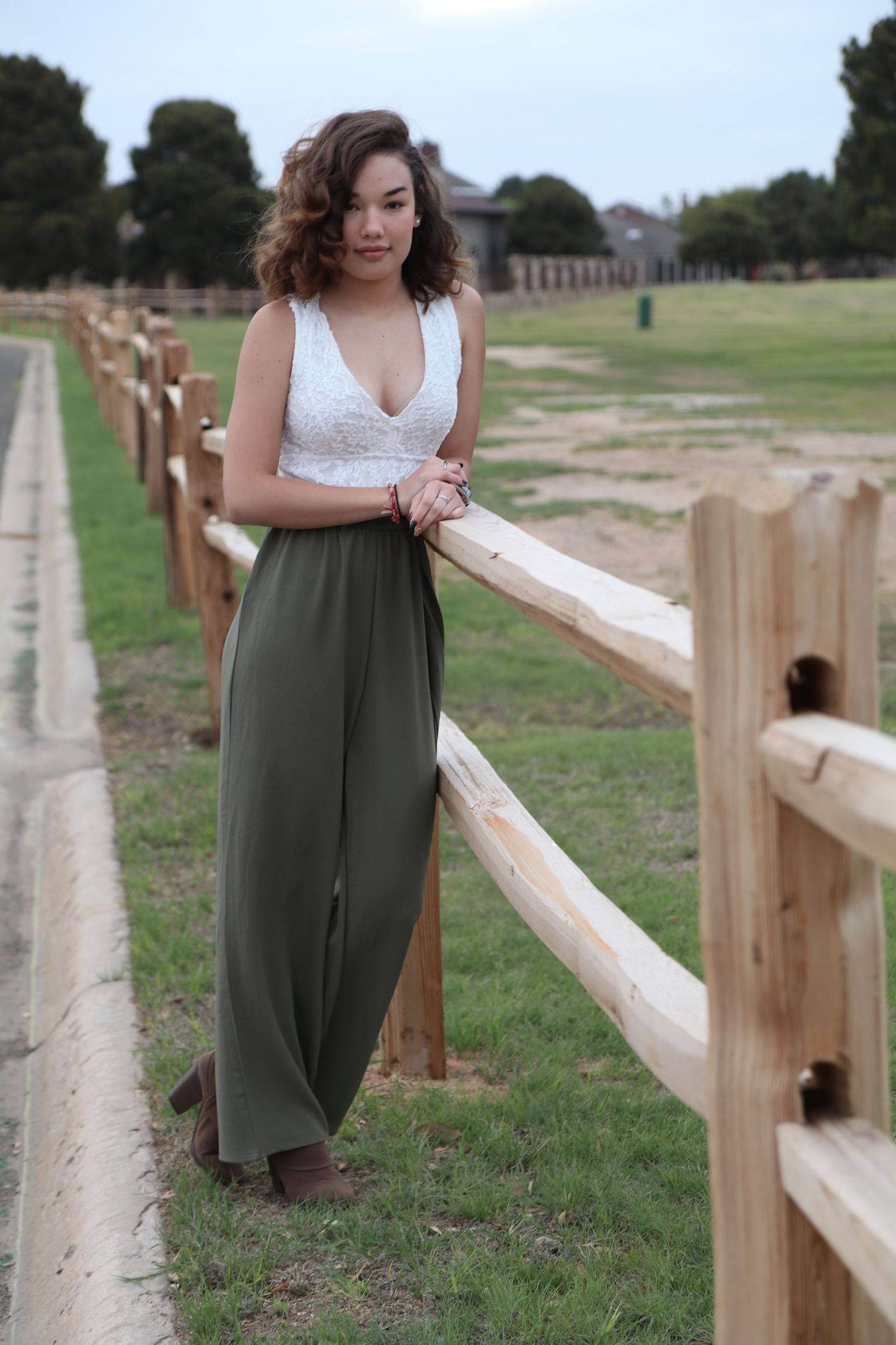 Woman in white top and olive palazzo pants leaning on wooden fence at rural location.