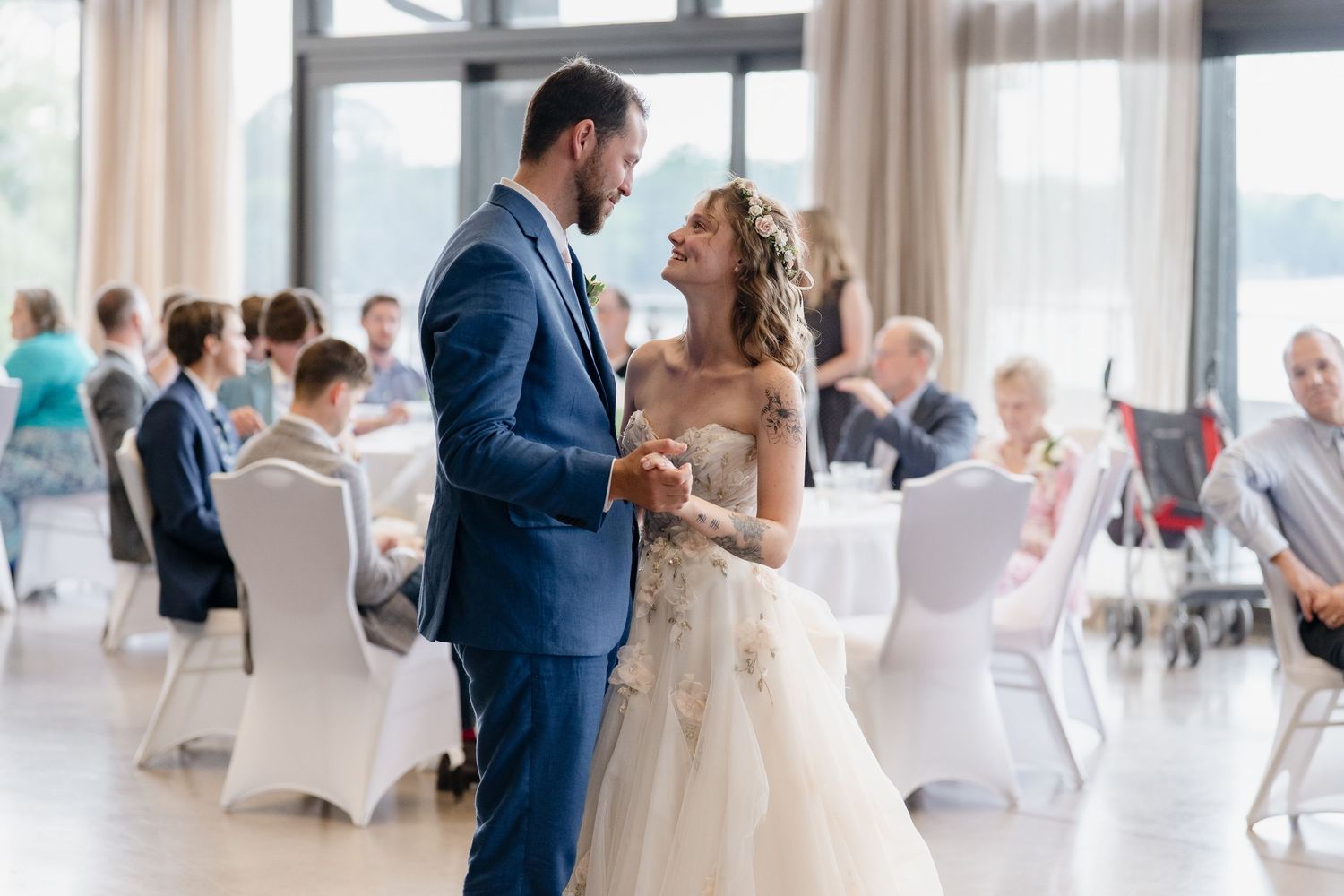 Wedding guests dance and celebrate during a reception in a bright, modern venue with white chairs and large windows.