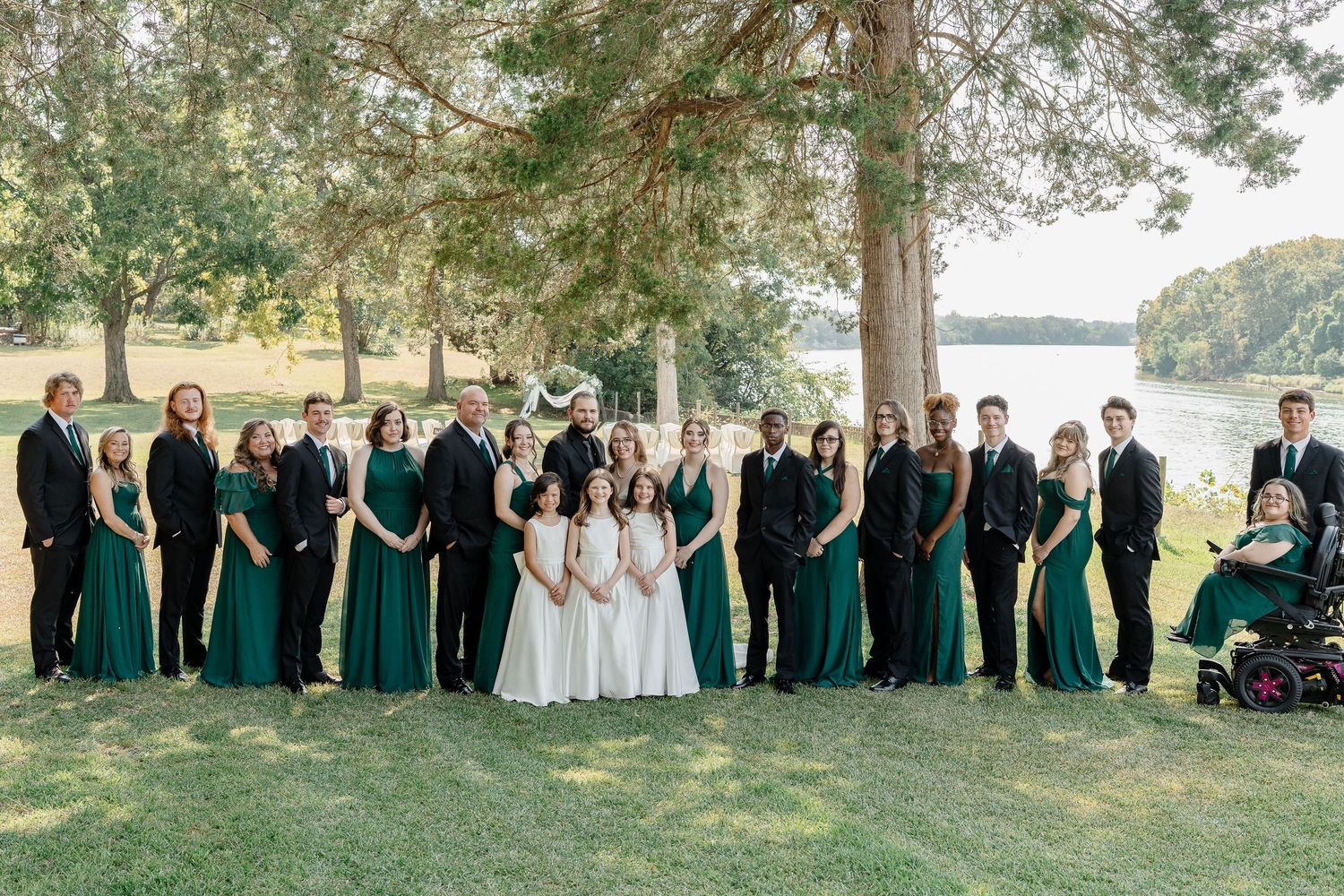 Large wedding party in emerald green dresses and formal attire poses together outdoors under tall trees near a lake.