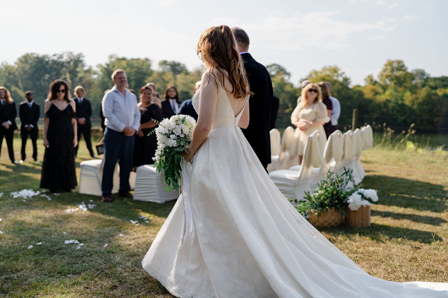A wedding celebration in a sunny outdoor setting with guests gathered around the bride and groom during the ceremony.