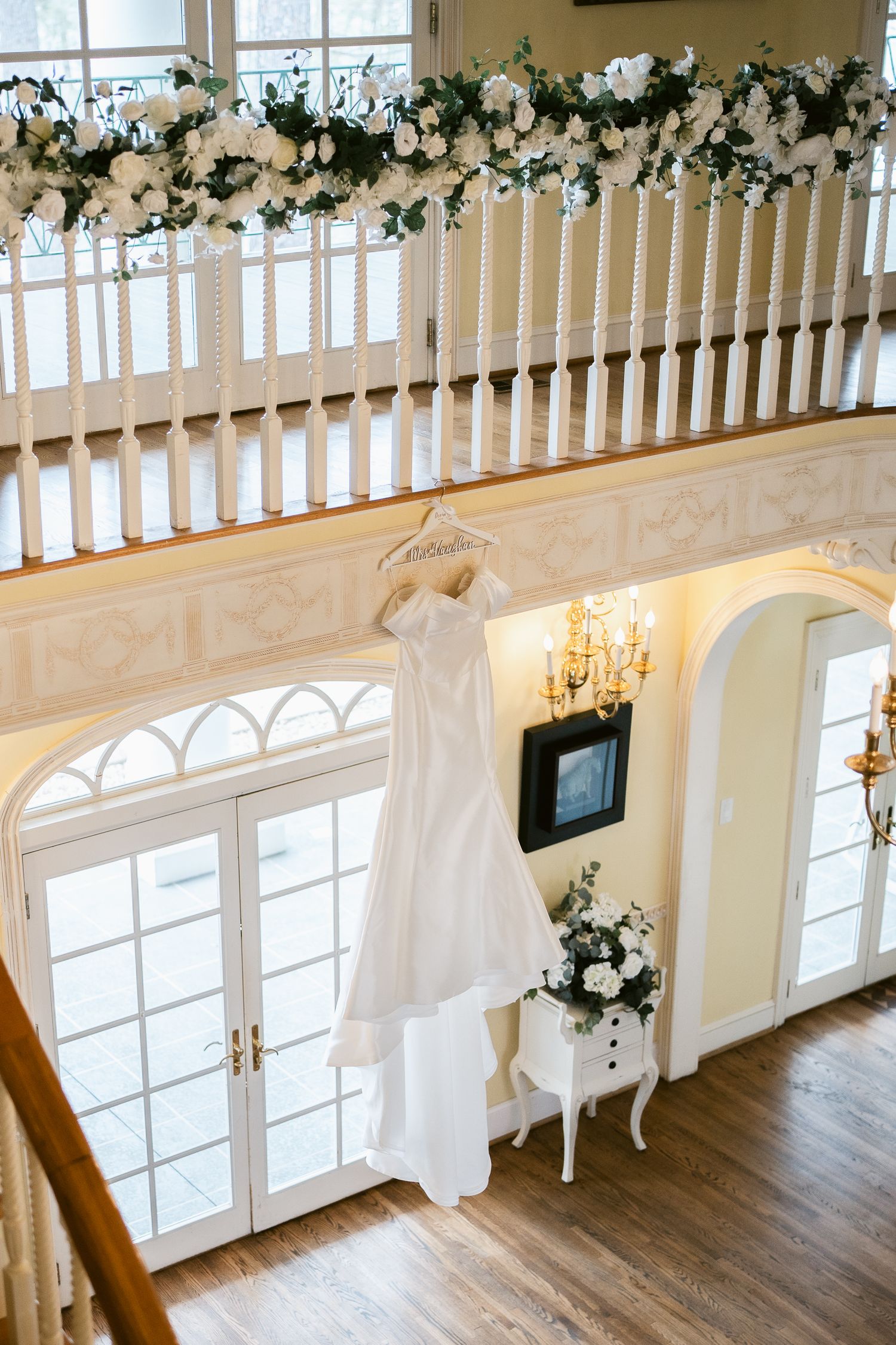 White wedding dress hanging by French doors with floral garland and chandelier decoration.