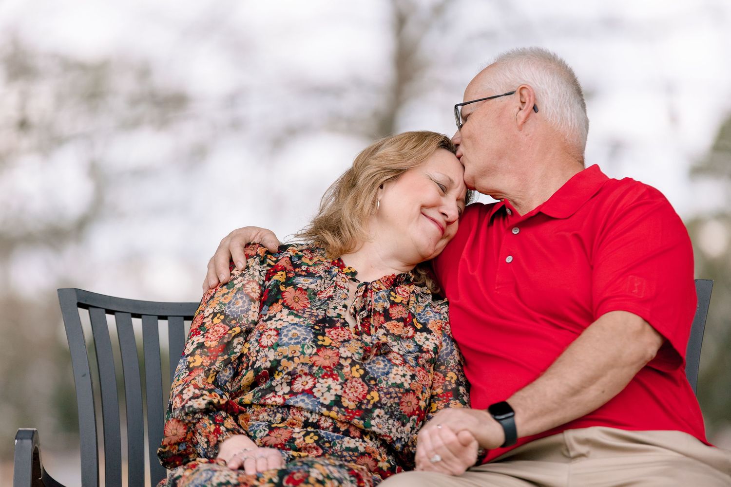 An elderly couple sits on a bench, sharing a tender romantic moment outdoors.