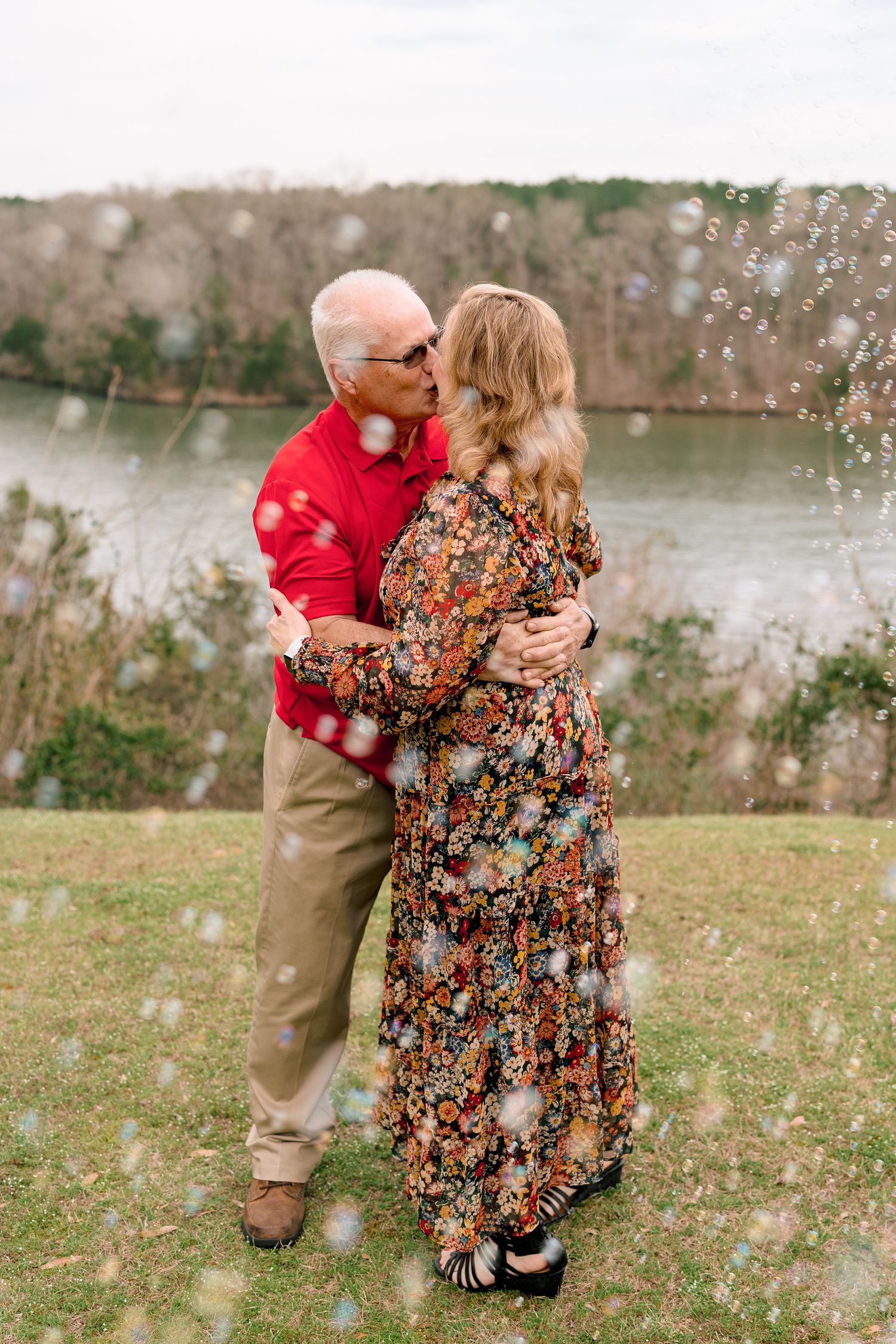 Older couple kissing and embracing outdoors near a river surrounded by floating bubbles.