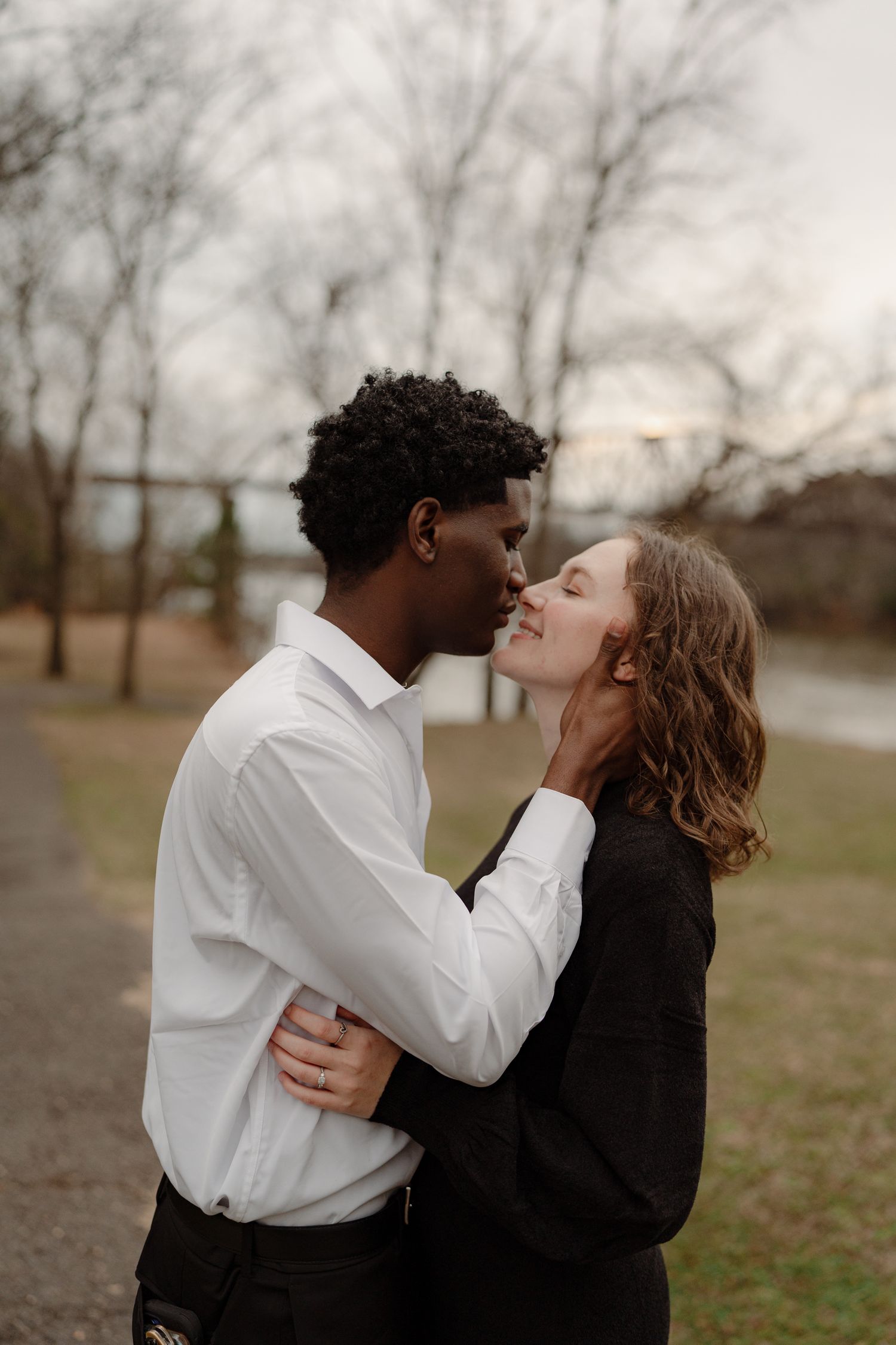 Romantic interracial couple sharing a tender kiss outdoors in a park with bare winter trees behind them.