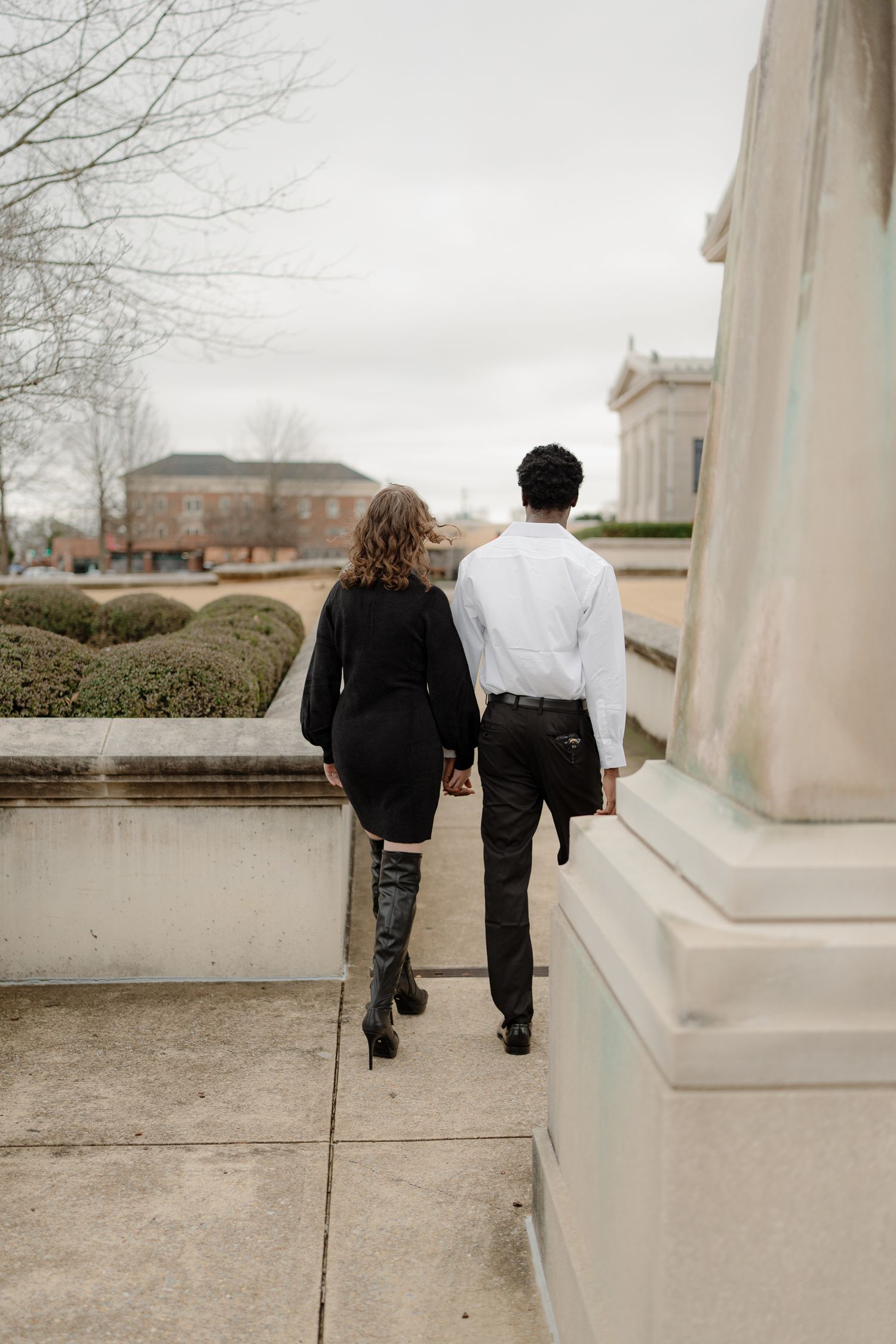 Interracial couple holding hands walking along a garden path, woman in black dress and boots, man in white shirt.