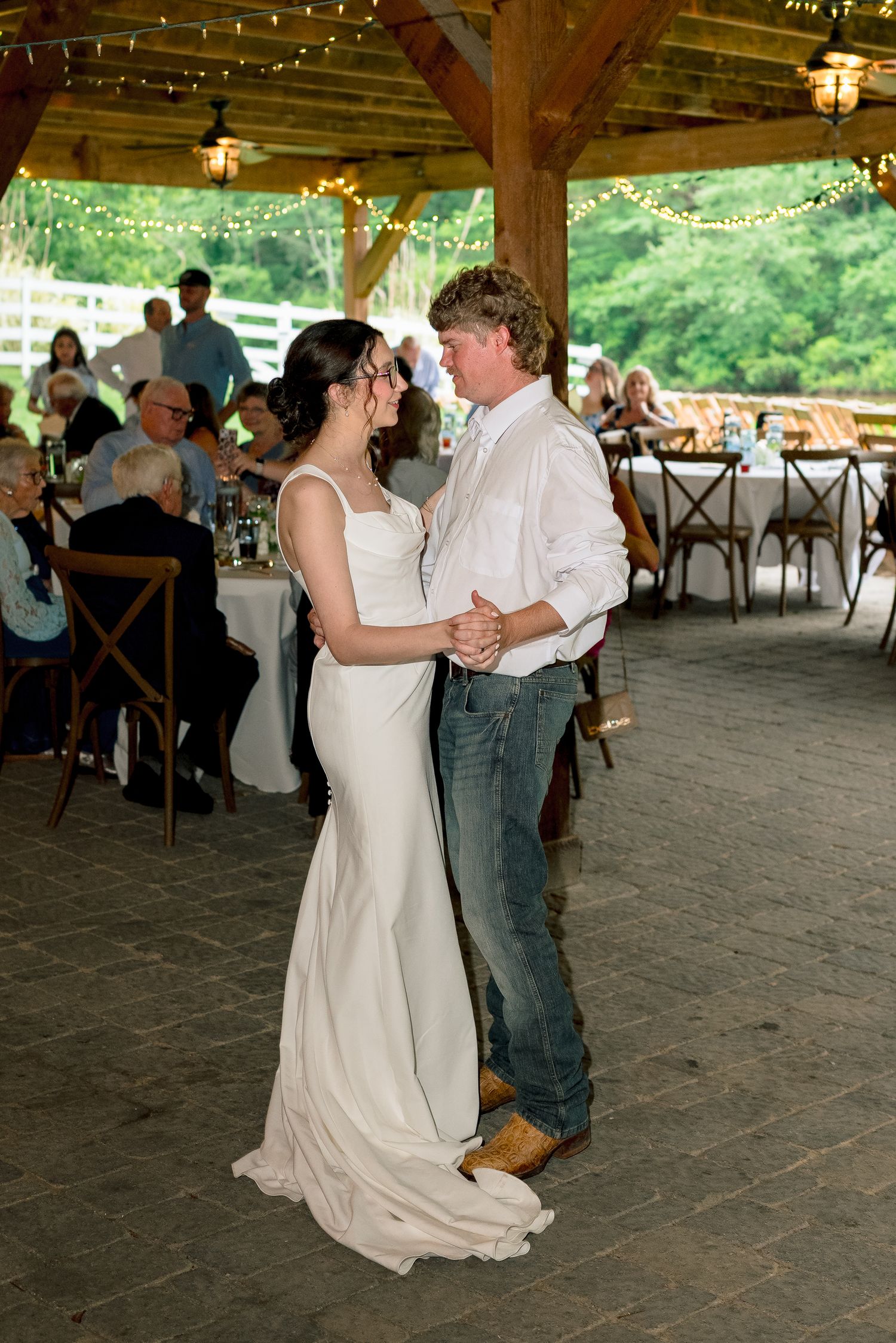 Bride and groom share first dance under string lights at rustic outdoor wedding reception pavilion.