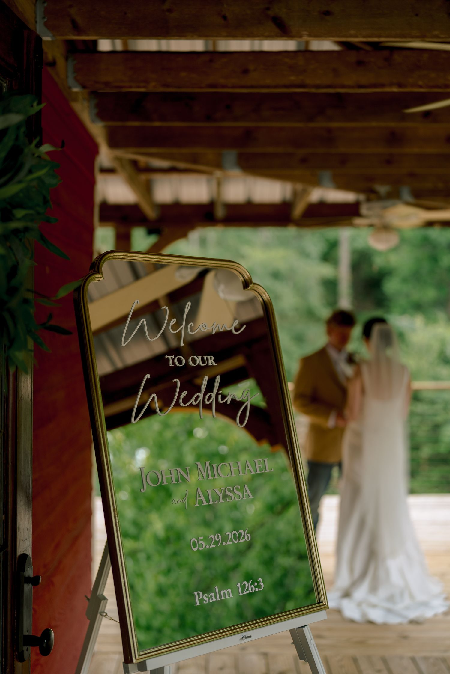 Elegant mirror wedding welcome sign for John Michael and Alyssa dated 05.29.2026 with couple in background.