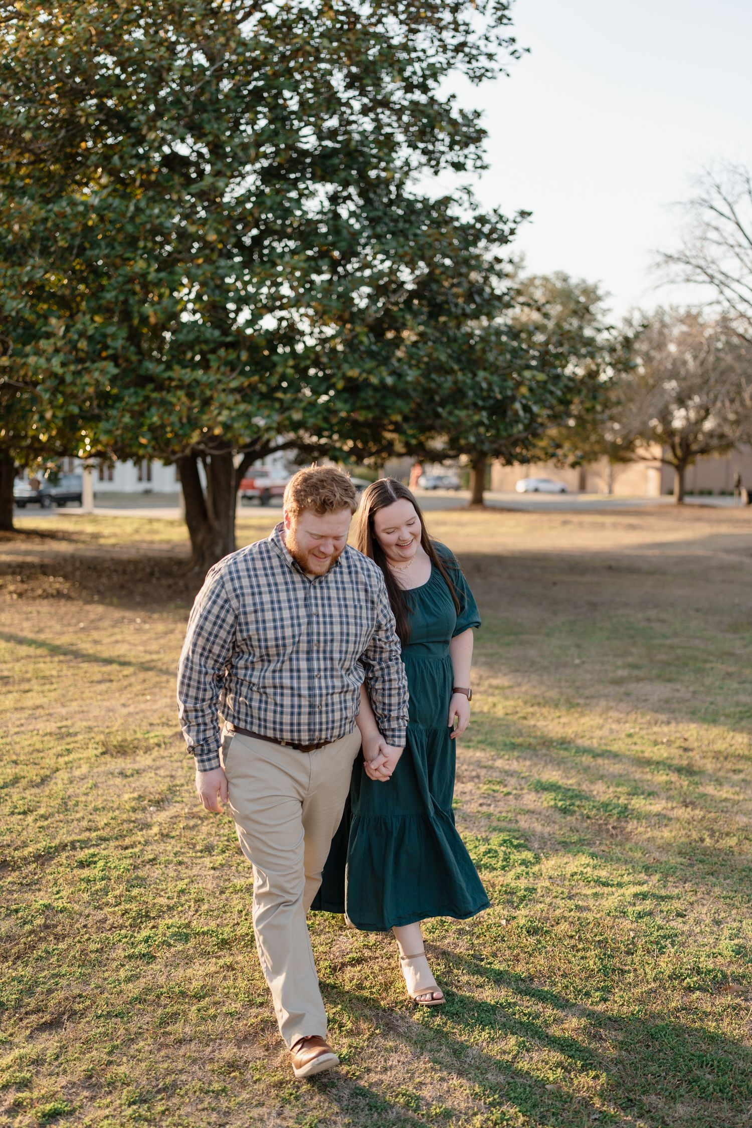 Couple holding hands walking through a sunny park with lush green trees during a golden hour engagement session.