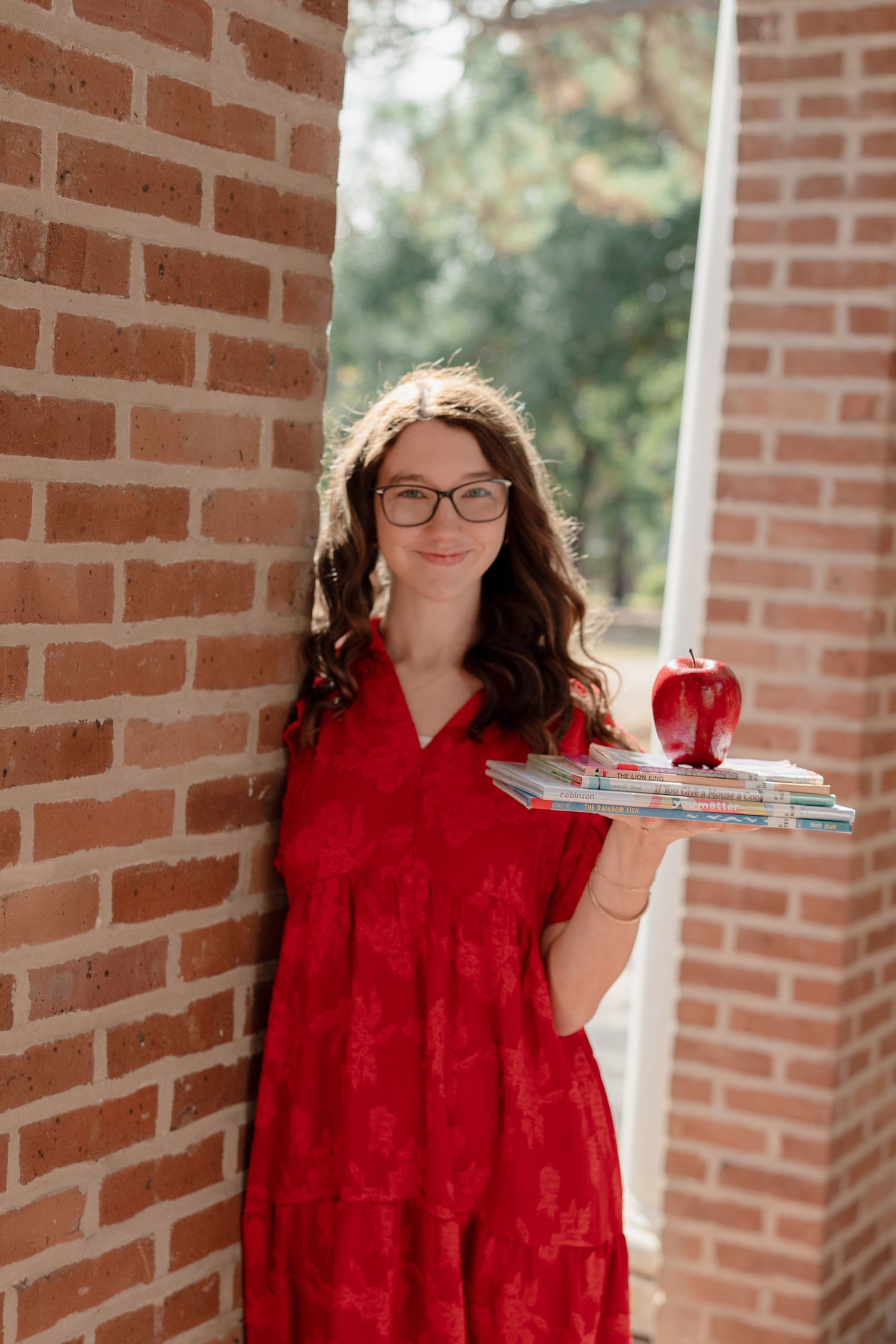 Person in red dress and glasses stands against brick wall with books and coffee cup.