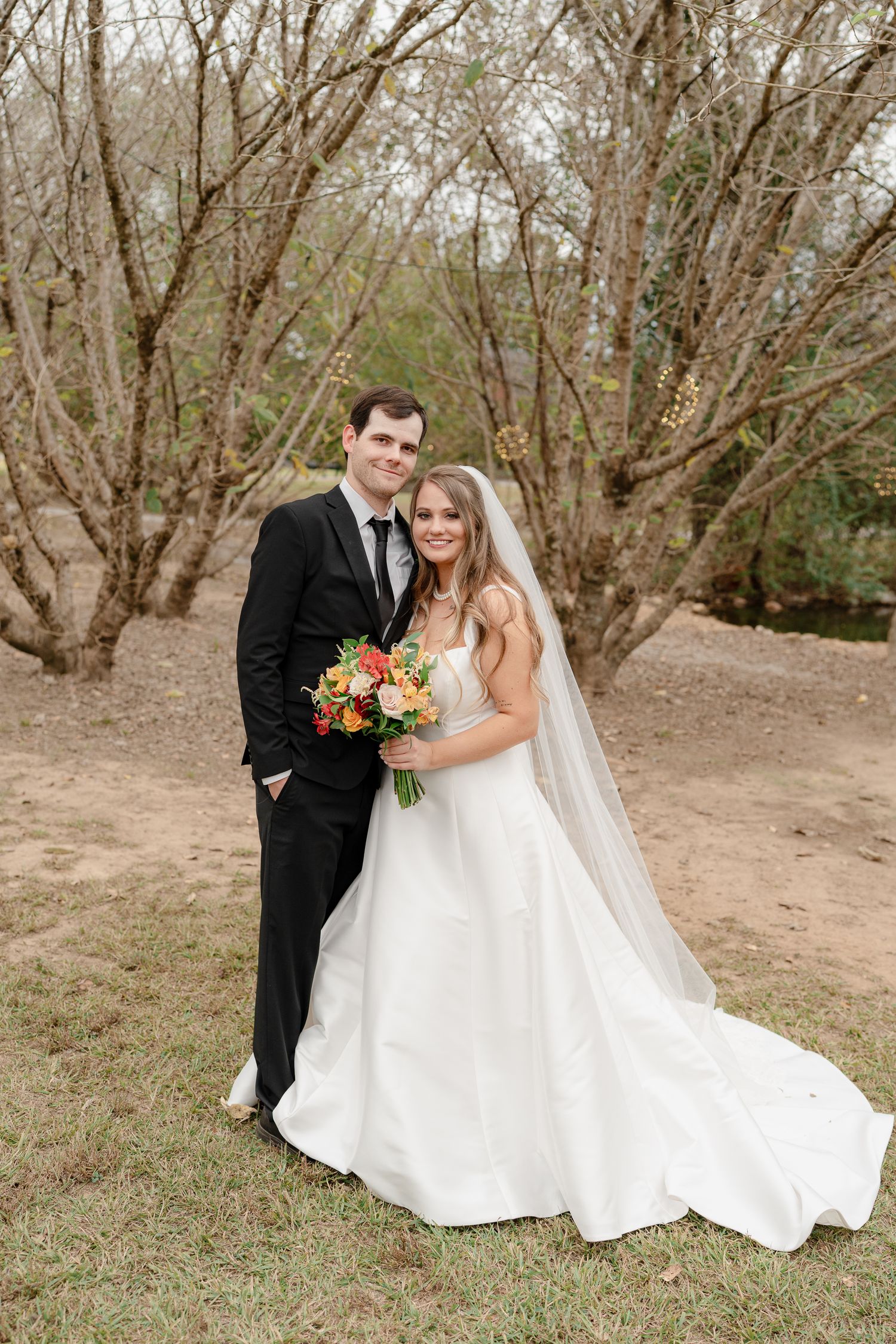 A bride and groom pose outdoors for a wedding photo among bare trees while holding a vibrant fall bouquet.
