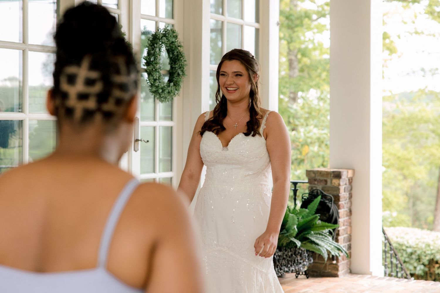Bride in lace wedding gown smiling during first look moment at elegant venue with greenery backdrop.