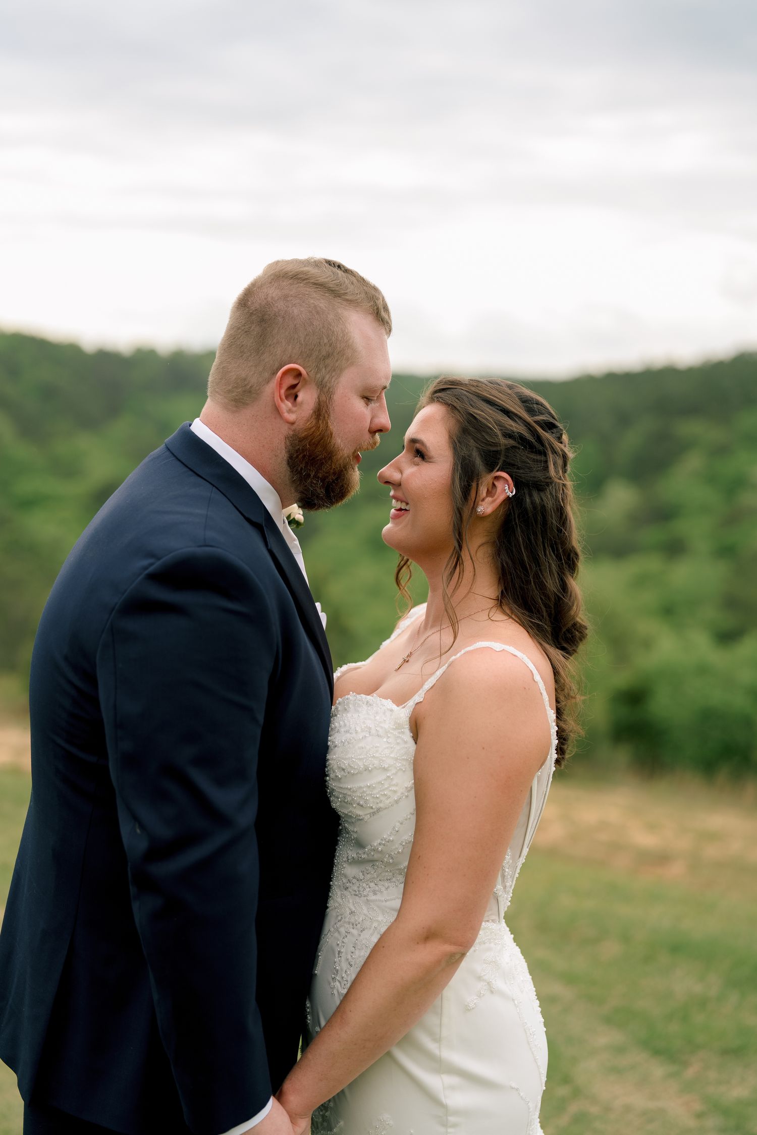 Bearded groom in navy suit sharing intimate romantic moment with smiling bride in lace wedding gown.