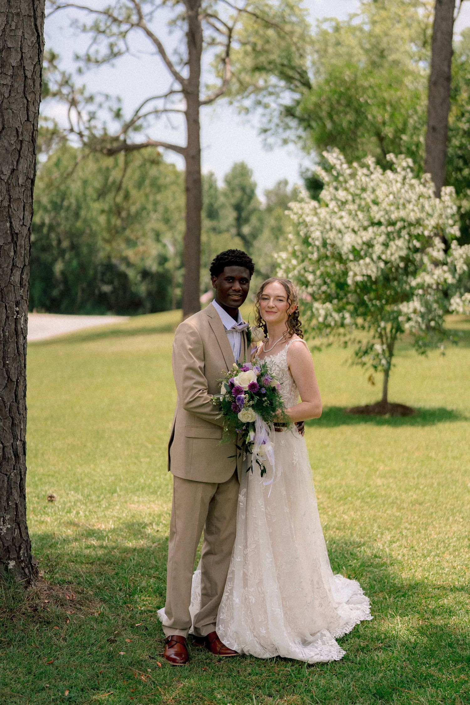 Interracial couple poses outdoors on wedding day, groom in tan suit, bride in lace gown holding purple bouquet.
