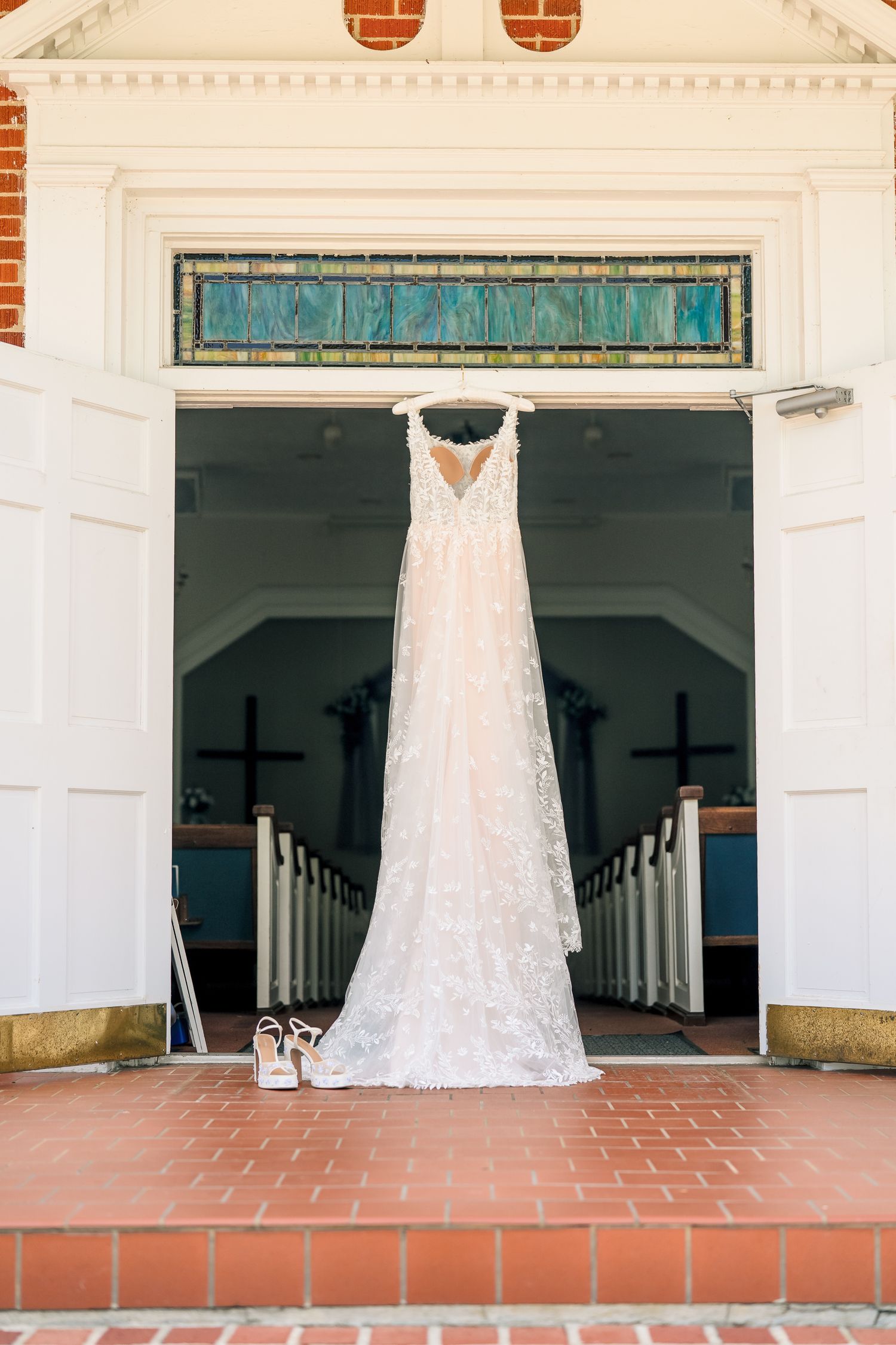 Elegant lace wedding gown hanging in church doorway with stained glass above and bridal heels on brick steps below.
