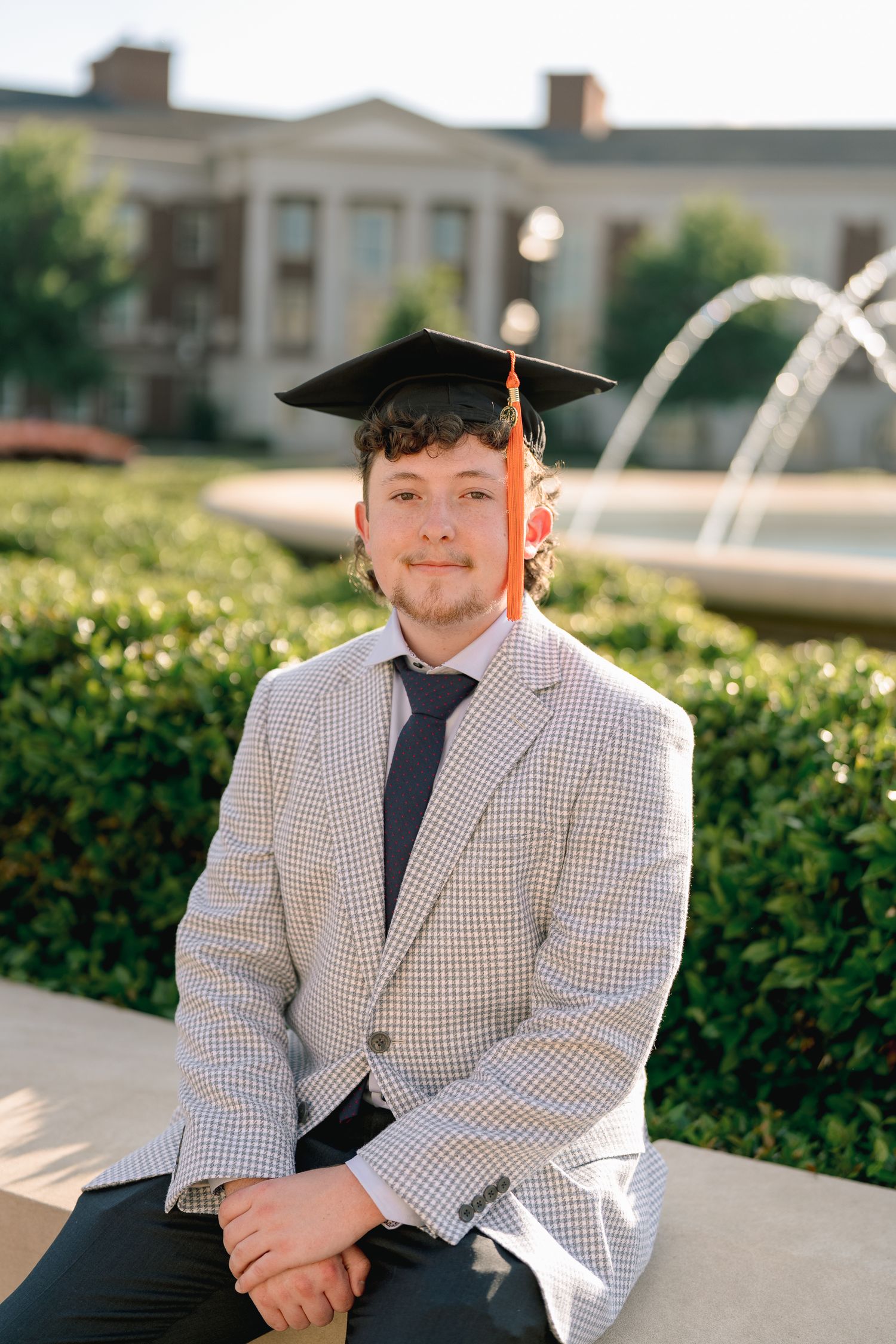 Graduate in checkered blazer and black mortarboard cap sits near a campus fountain smiling.