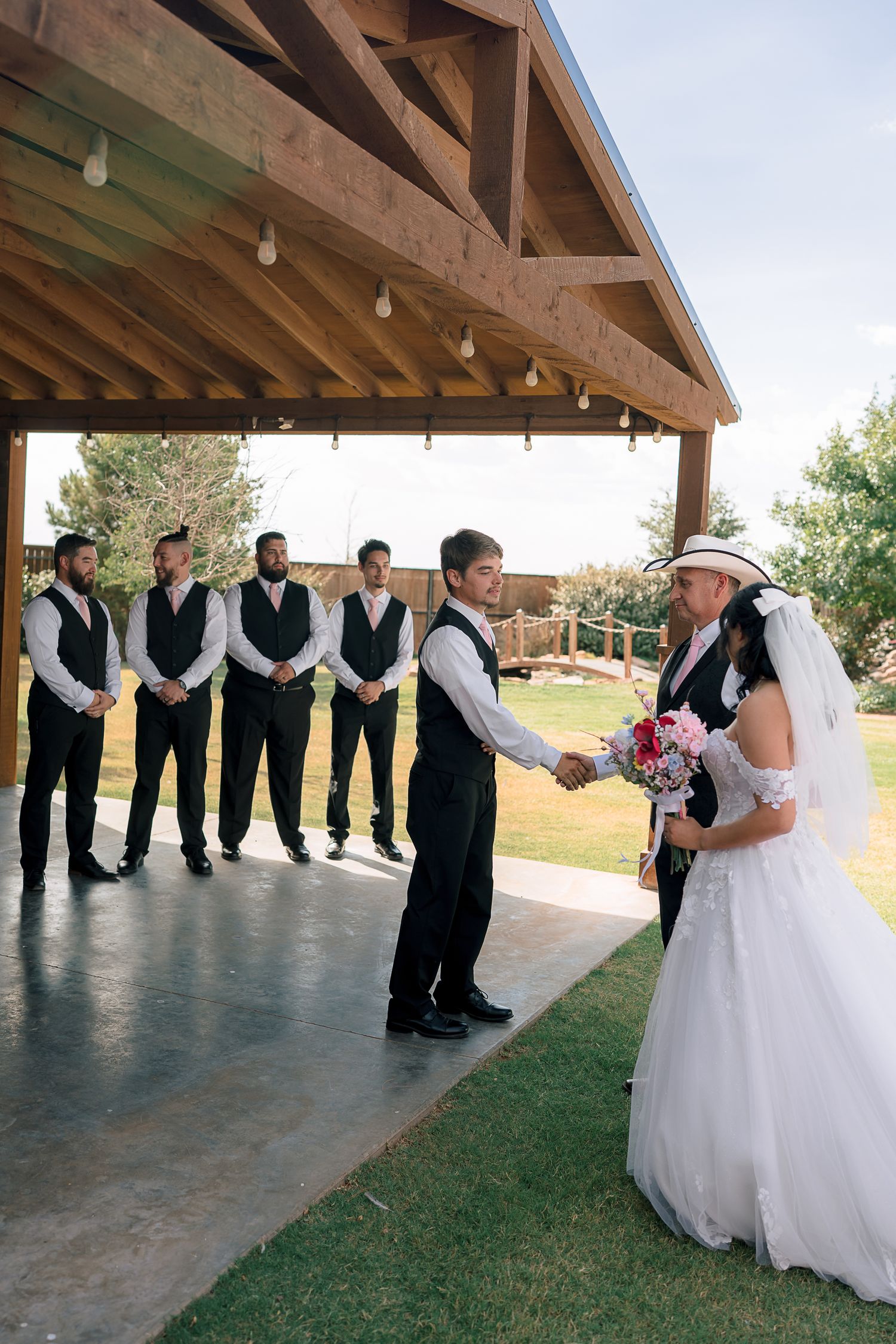 Groom greets bride and her wedding party under a wooden outdoor ceremony pavilion with groomsmen lined up.