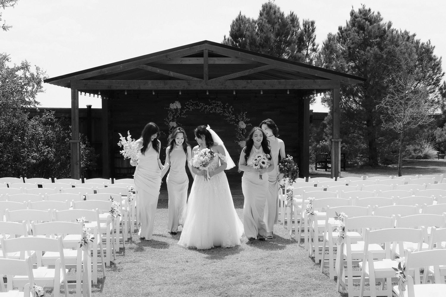 Black and white photo of bride and bridesmaids walking toward outdoor wedding ceremony pavilion.