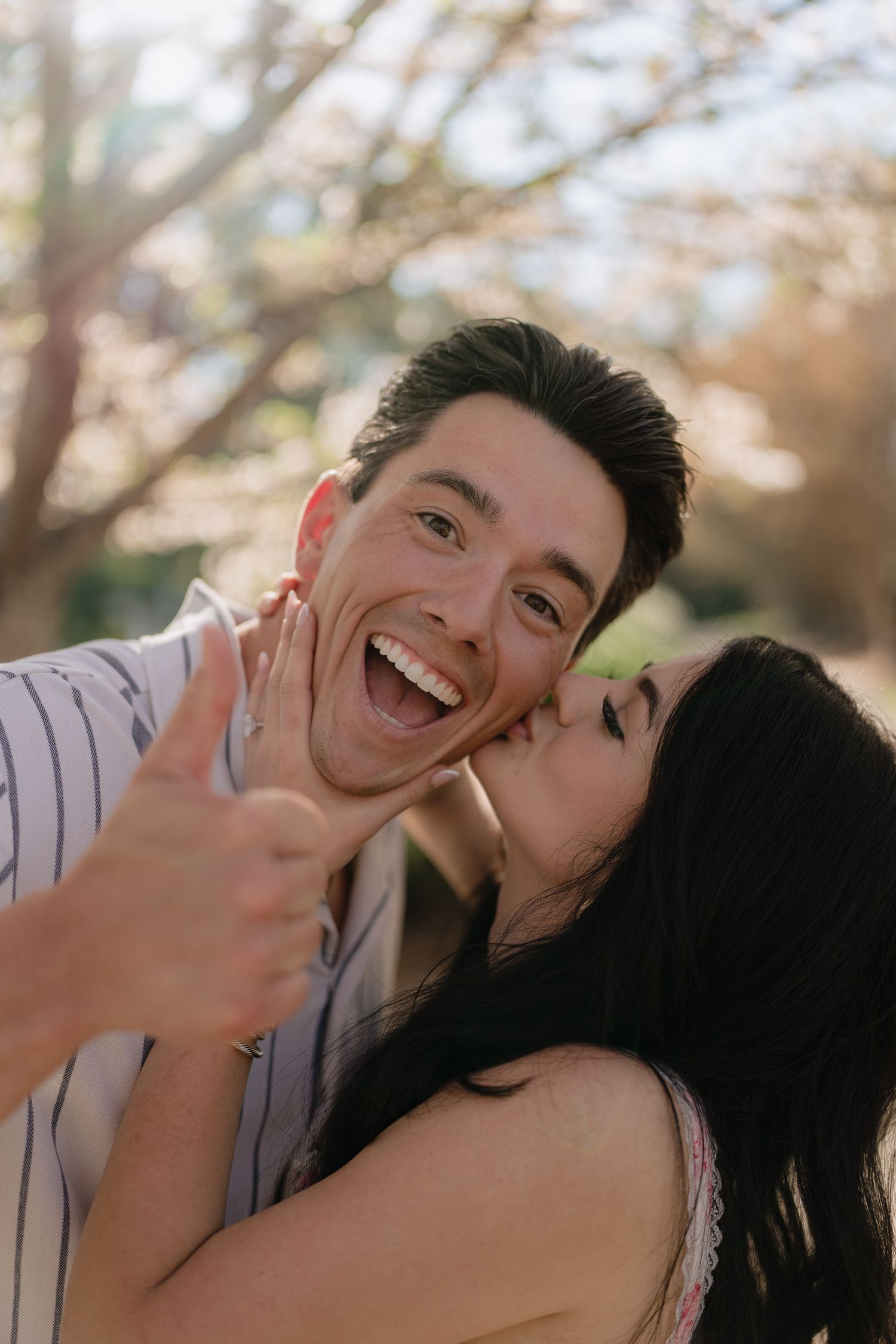 Happy couple sharing a romantic kiss outdoors, man giving thumbs up near blooming cherry blossom trees.