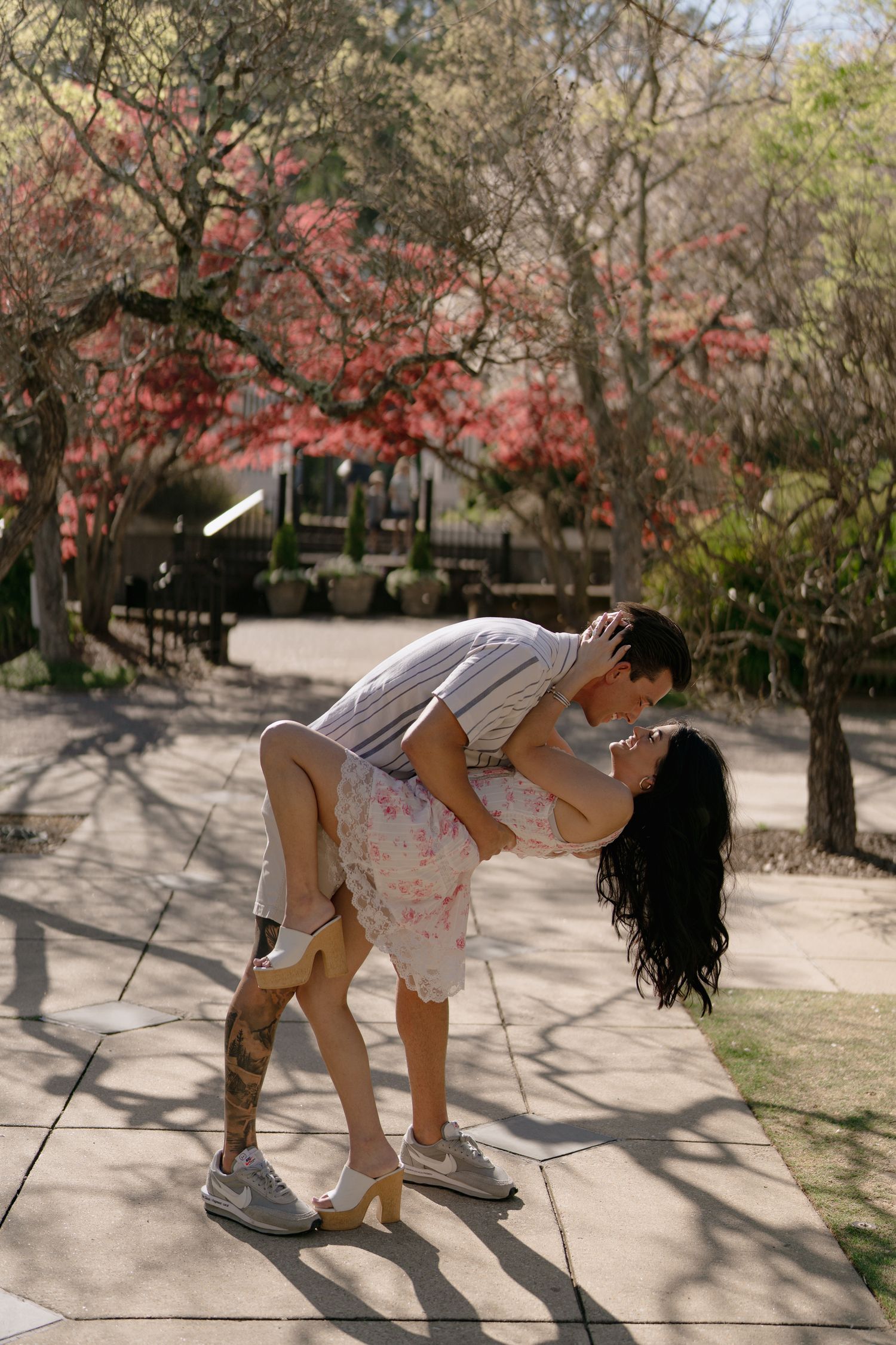 Couple sharing a romantic dip kiss on a sunny garden path surrounded by blooming red and pink trees.