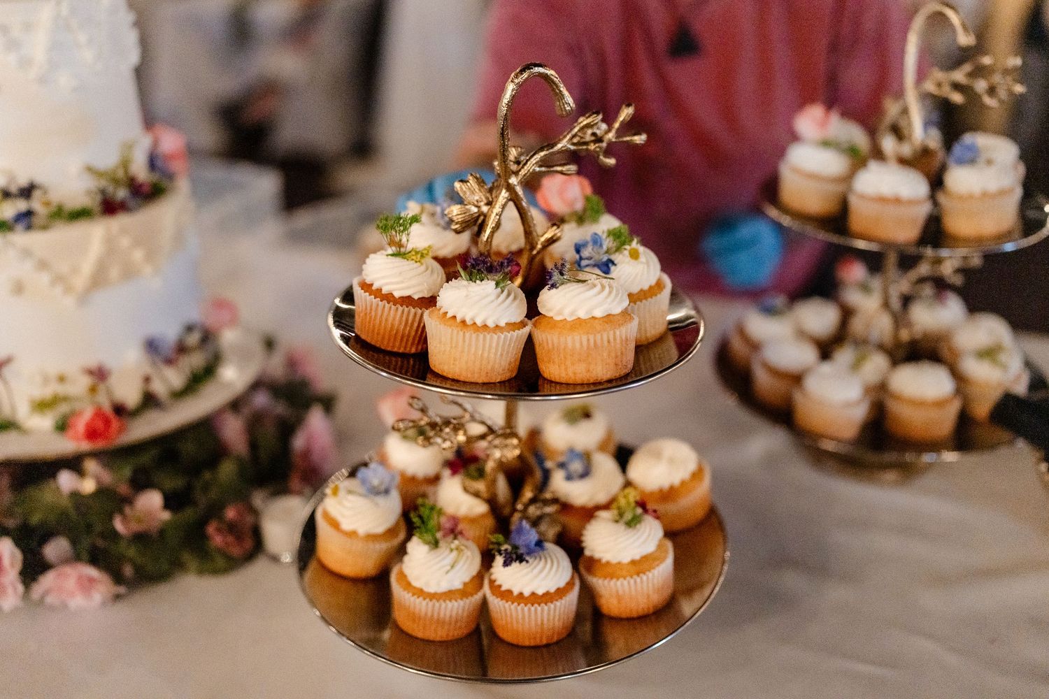 Elegant tiered display stands filled with frosted cupcakes decorated with flowers and greenery at a wedding reception.