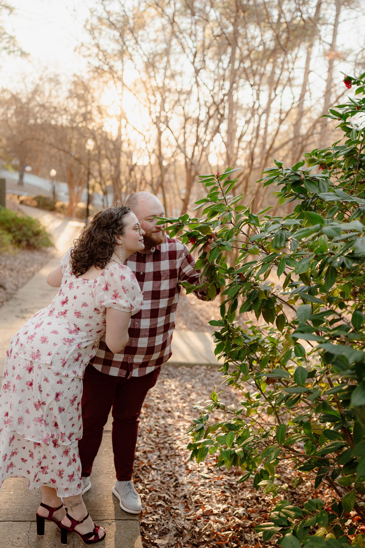 A couple in floral dress and plaid shirt share playful moments among autumn trees at sunset.