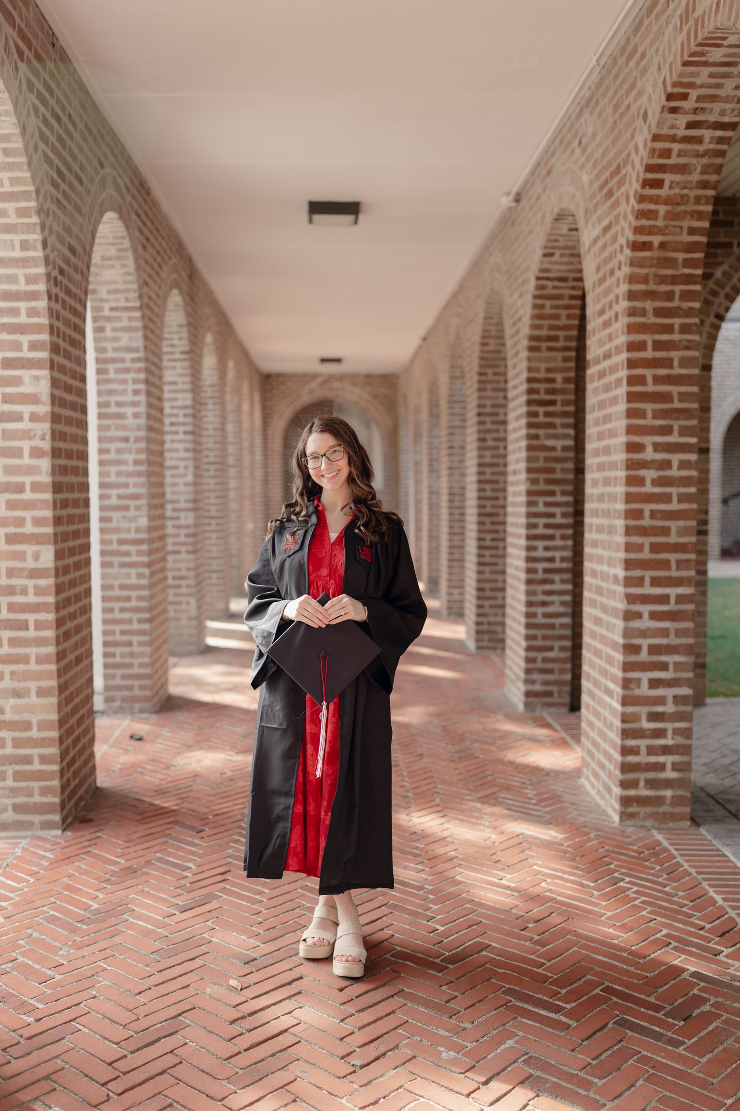 A person in black and red attire stands in a brick-lined colonnade with repeating arched columns and herringbone floor pattern.
