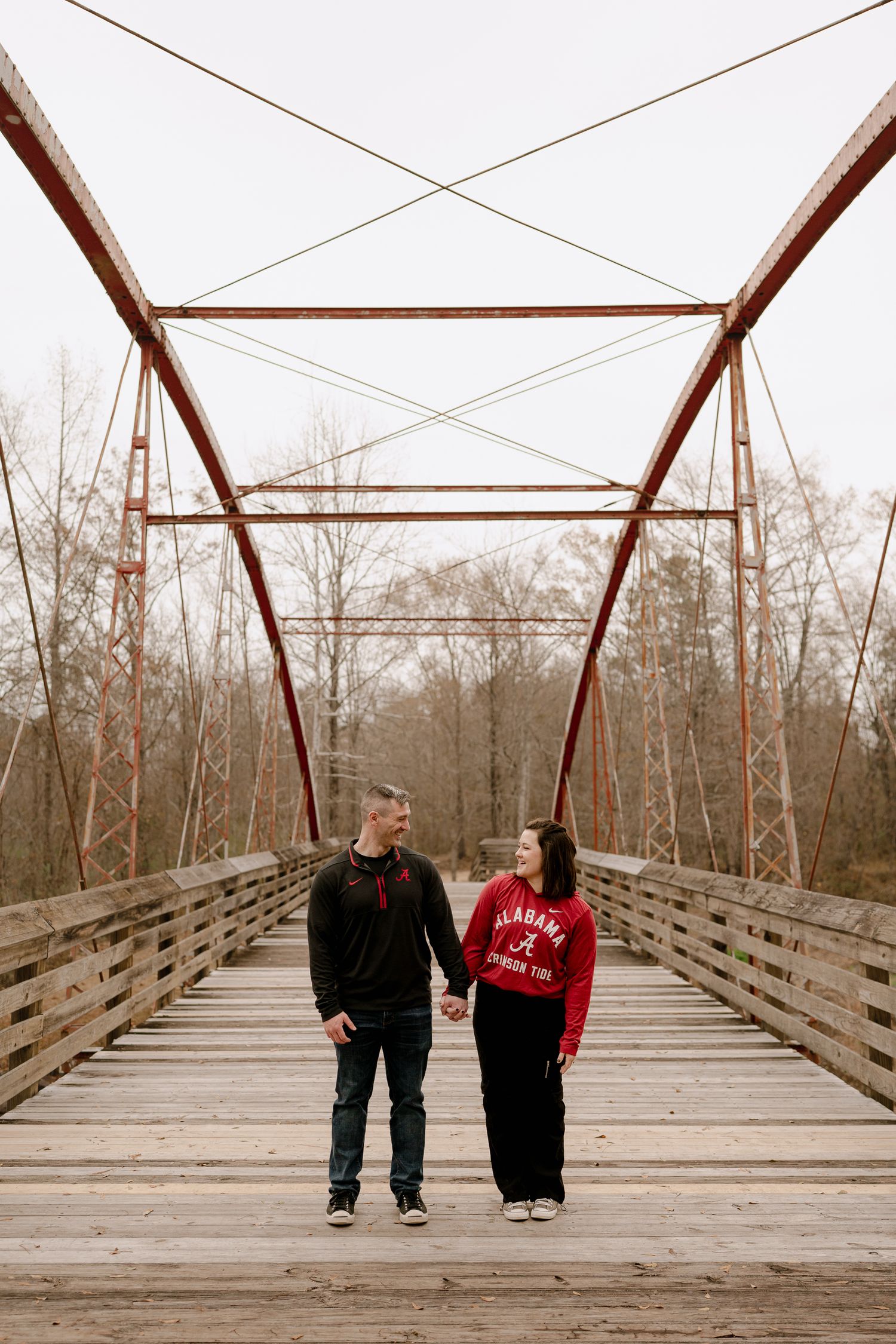 Two people walking and interacting on a rustic wooden bridge surrounded by bare trees in winter.