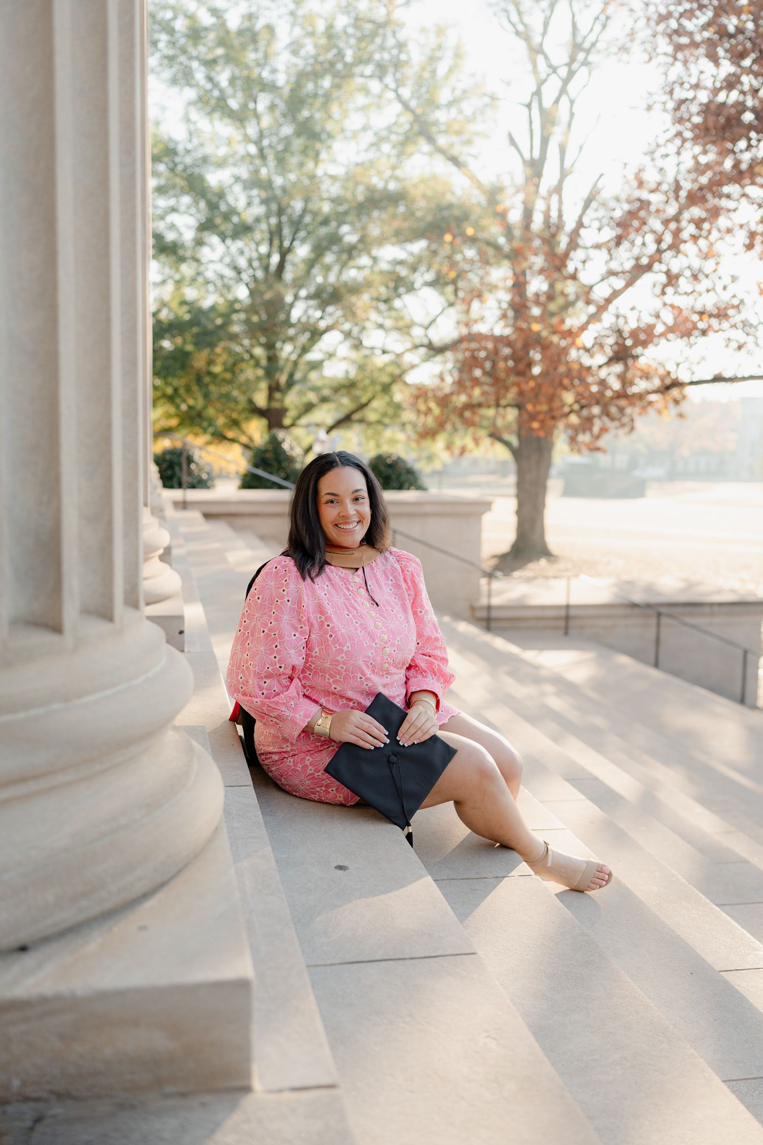 A graduate in pink dress sits casually between white columns during a sunny graduation photoshoot.