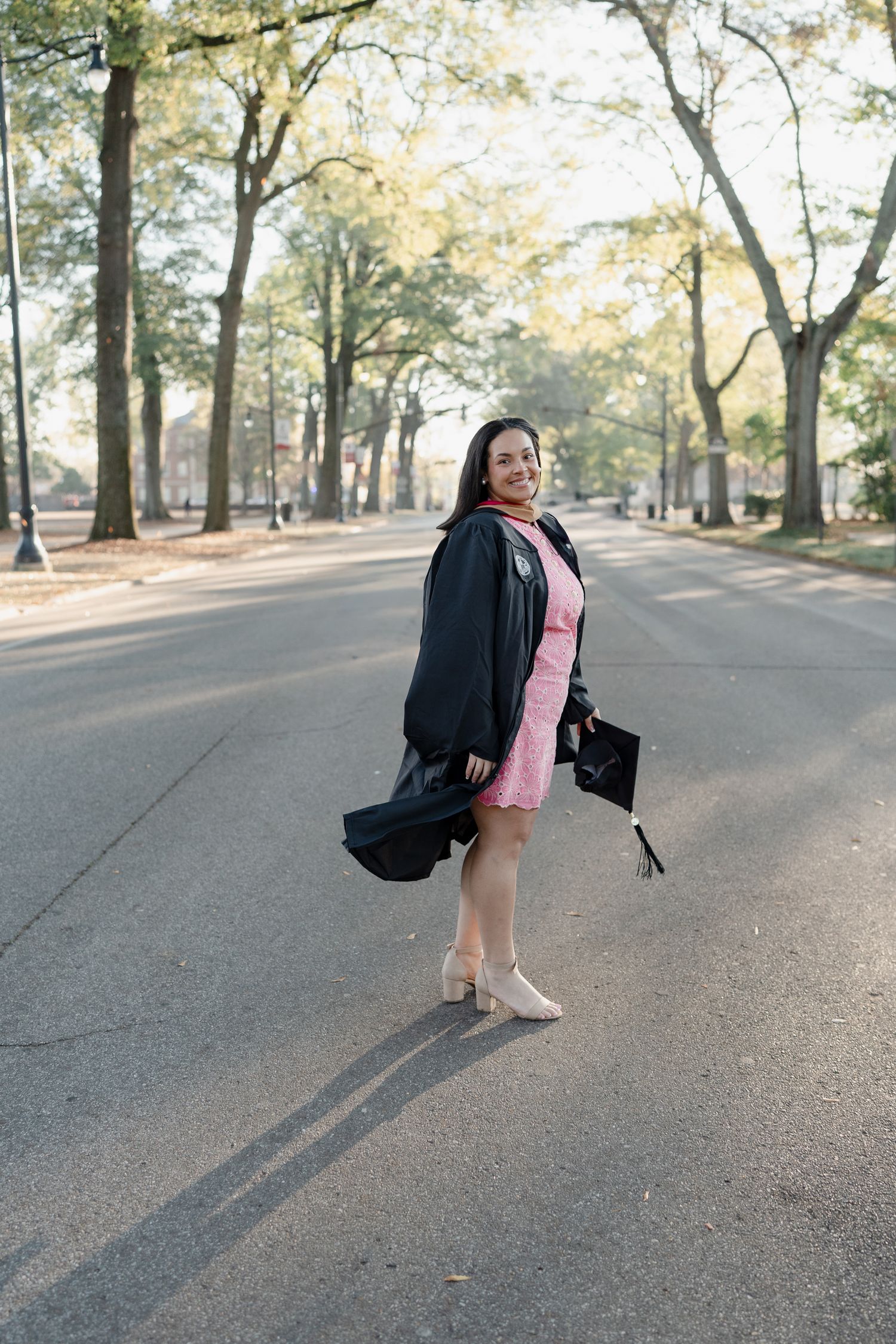 A graduate in a pink dress and black graduation gown poses on a tree-lined path during a sunny autumn day.