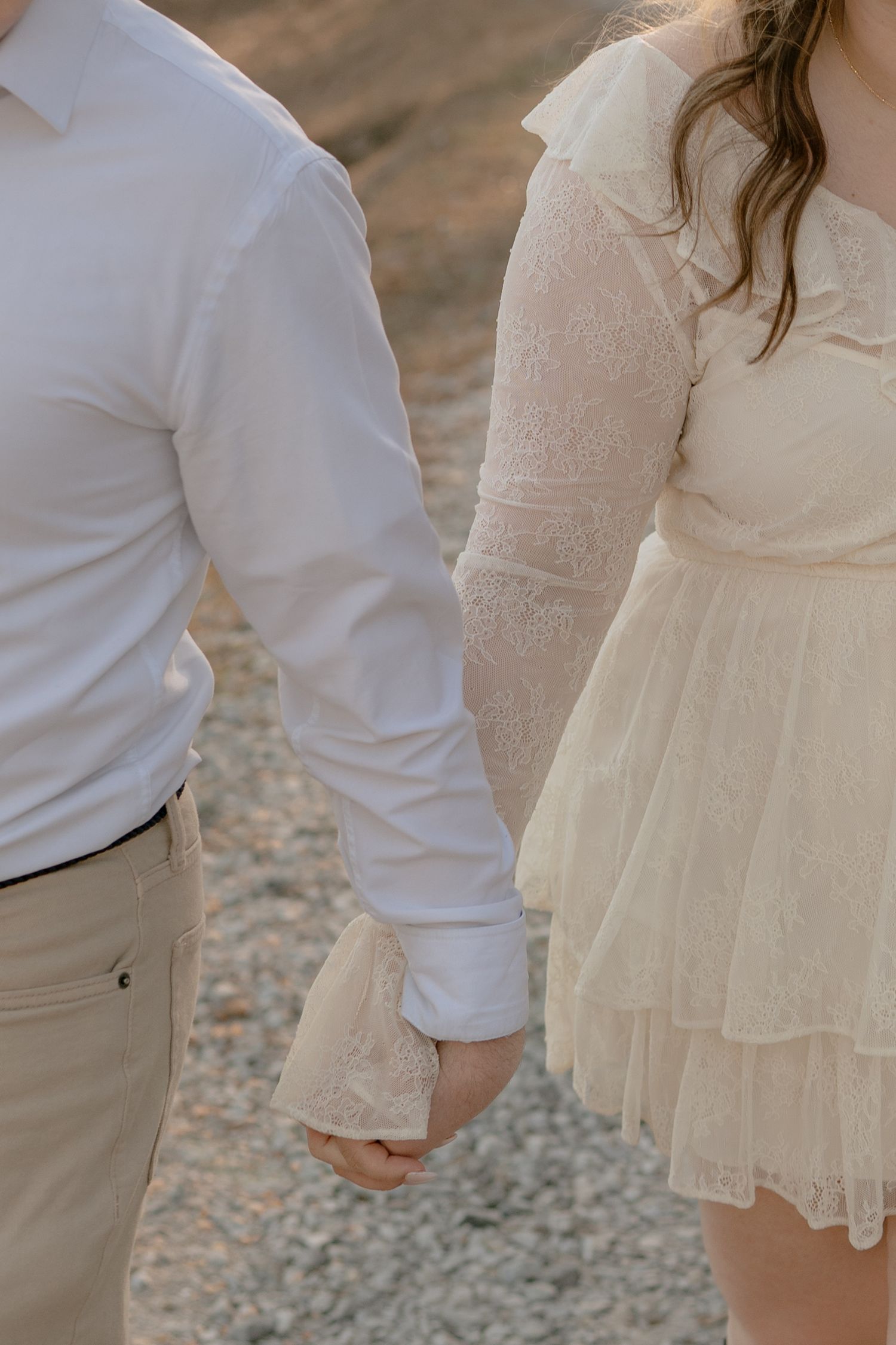 Close-up of couple holding hands, man in white shirt and woman in cream lace dress outdoors.