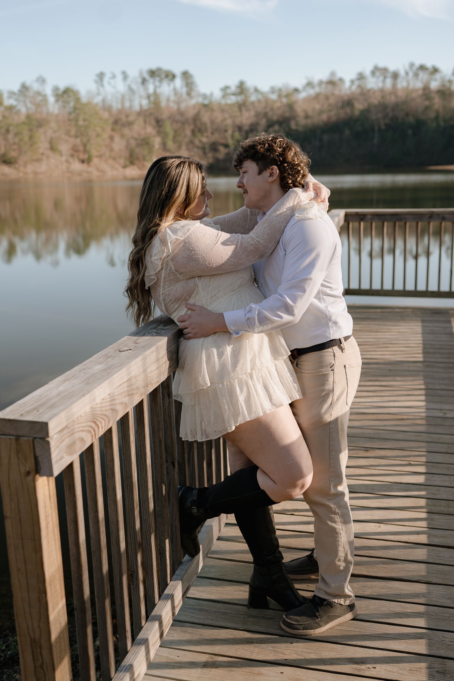 A couple embraces on a wooden dock by a serene lake, smiling at each other in warm sunlight.